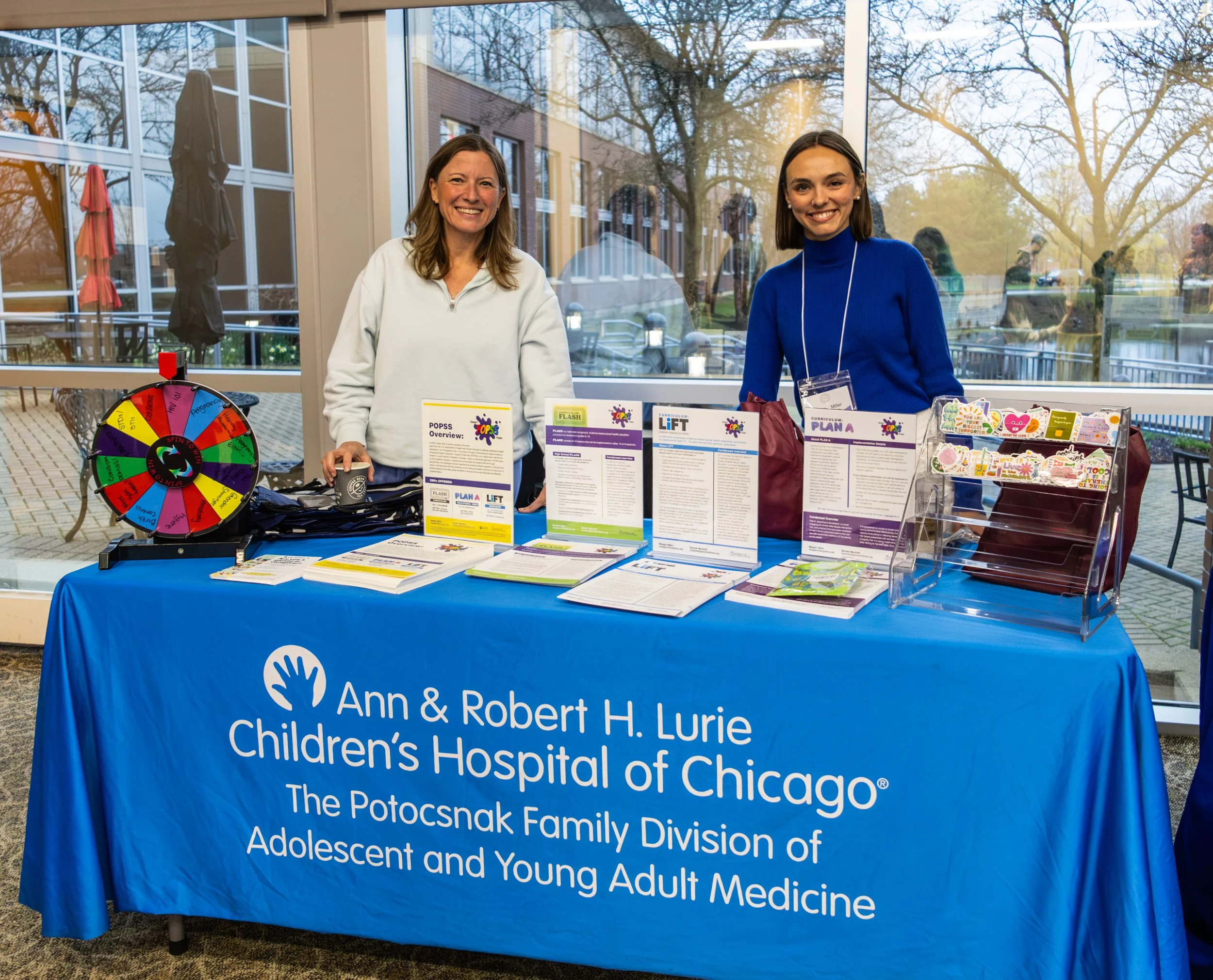 Two women standing behind a table at a community event, with informational pamphlets and a colorful prize wheel. The table has a blue tablecloth with the logo and name of Ann & Robert H. Lurie Children's Hospital of Chicago. The background shows large windows and trees outside.