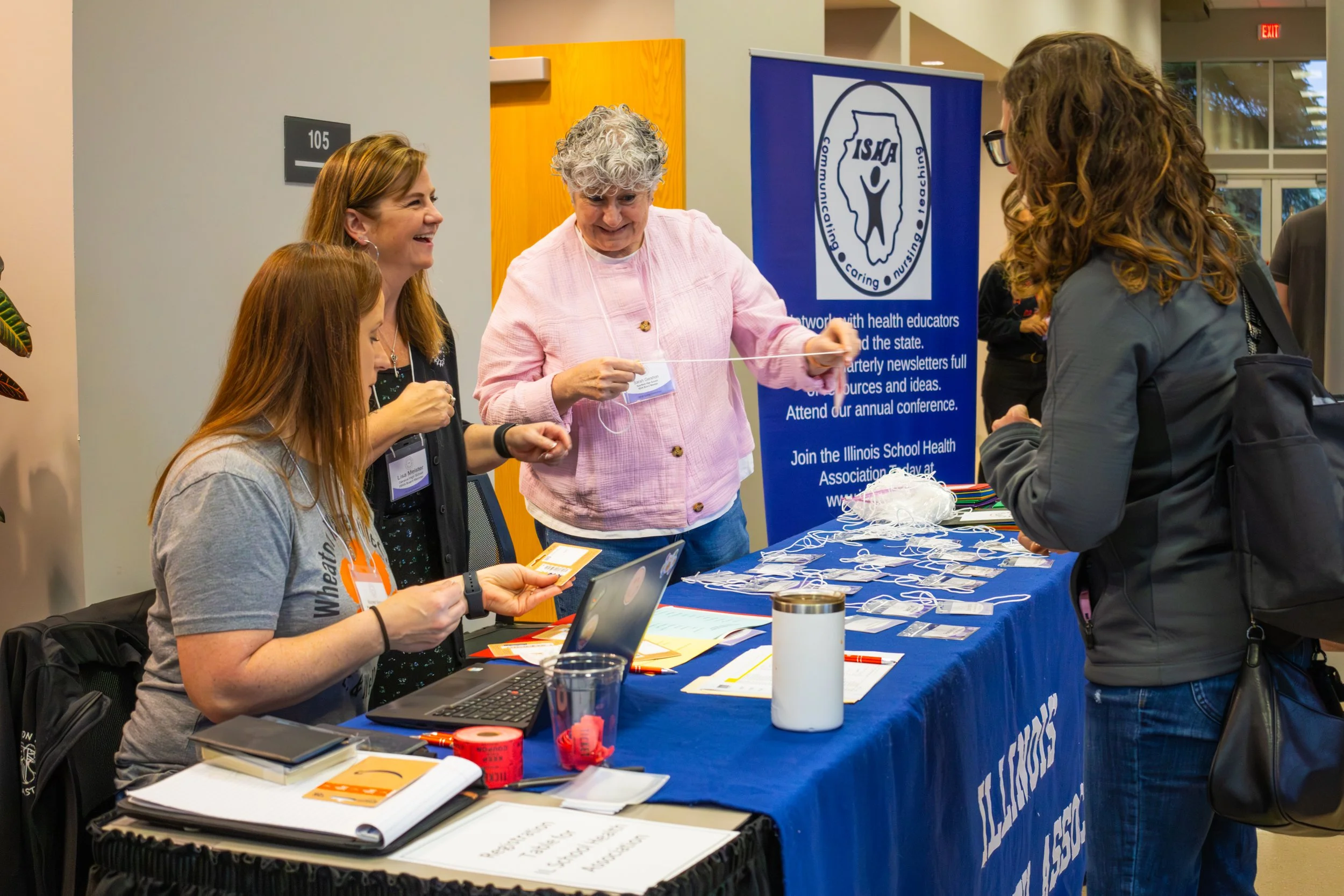 Four women at a conference table engaging in conversation. The table has various items including notebooks, a laptop, and name tags. Behind them is a blue banner with a logo and text about health education, and a window showing a building outside.