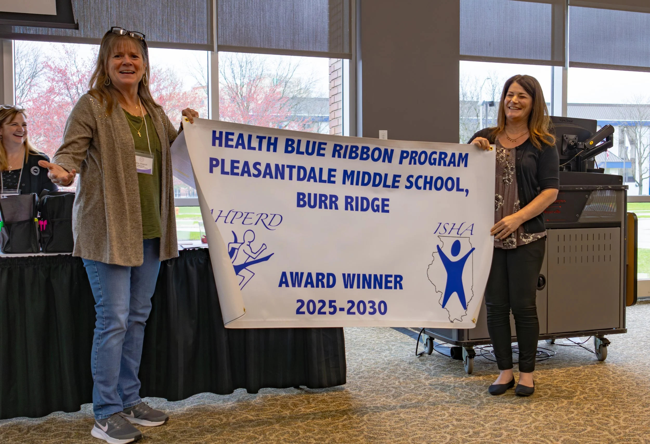 Two women holding a large banner at an indoor event. The banner reads 'Health Blue Ribbon Program Pleasantdale Middle School, Burr Ridge, Award Winner 2025-2030.' One woman on the left is wearing a beige cardigan and green shirt, while the woman on the right is dressed in black with a patterned dress underneath. They are smiling, and there is a third person partially visible on the far left. Large windows show trees outside, and the room has a black table and electronic equipment.