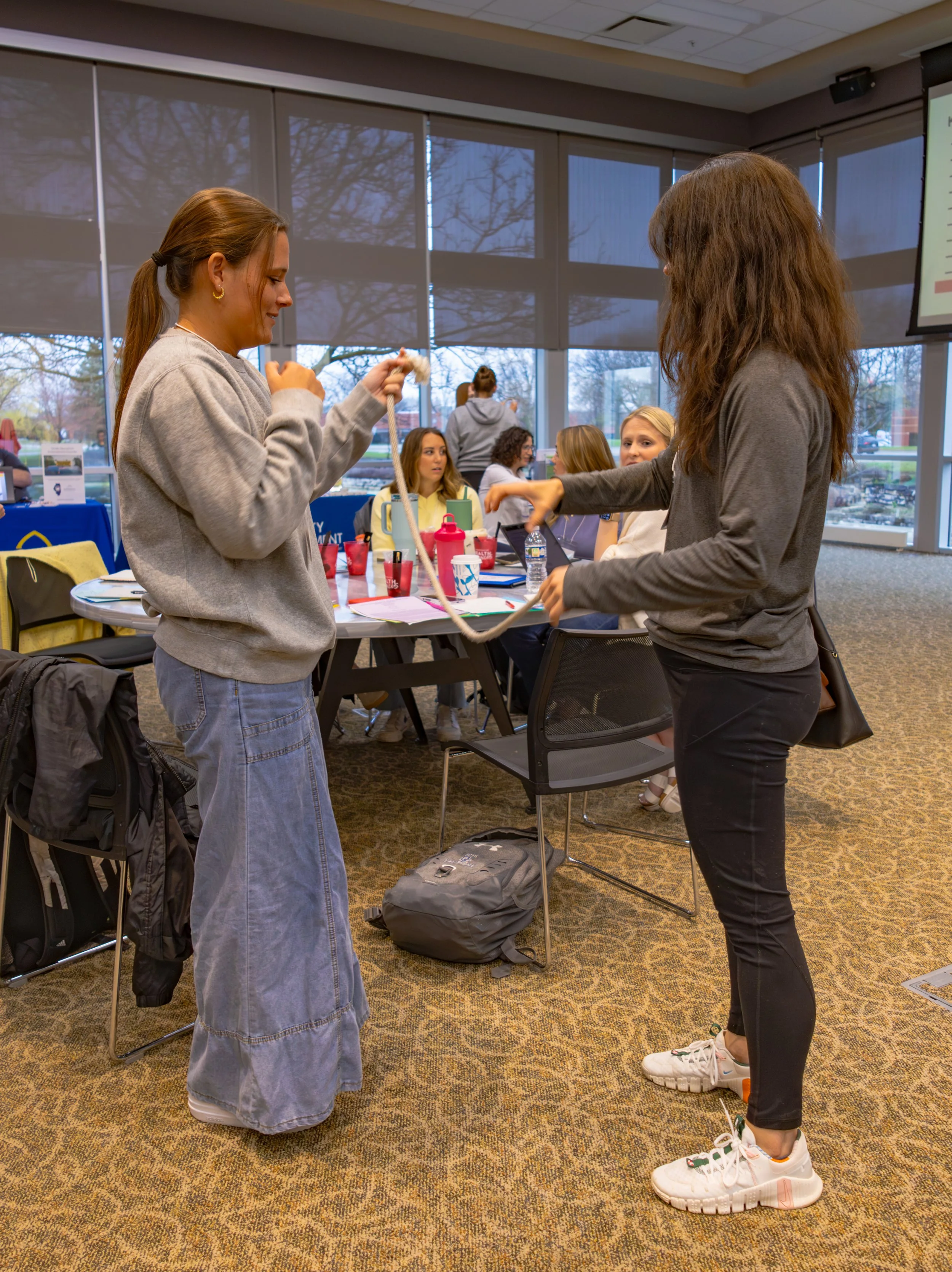 Two young women standing and holding a rope, engaging in a relationship-building activity during a workshop in a conference room. Several people are seated at a table in the background, working on laptops and talking. The room has large windows with blinds, and outside trees and a parking lot are visible.