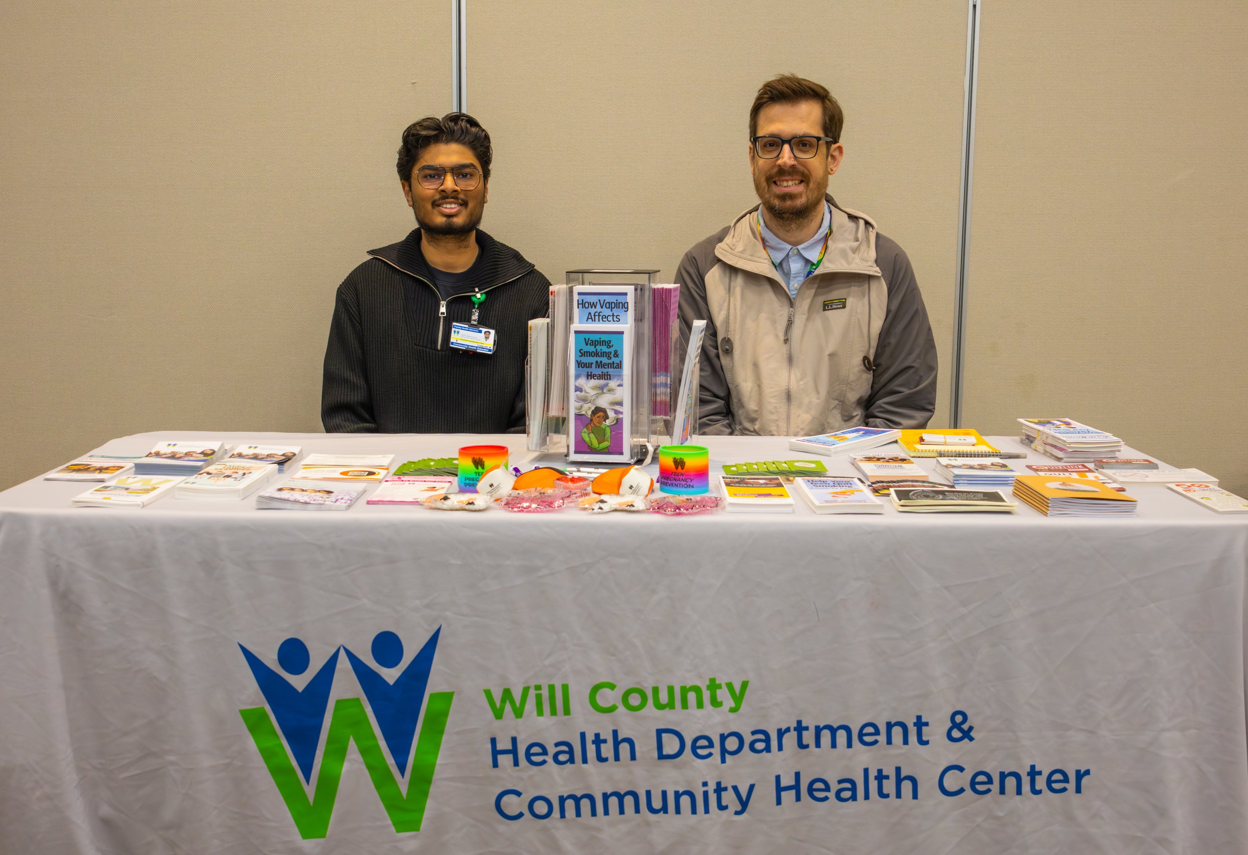 A table with educational flyers and pamphlets about vaping and health, with a clear sign promoting Will County Community Health Center, and two men standing behind the table in an indoor setting.