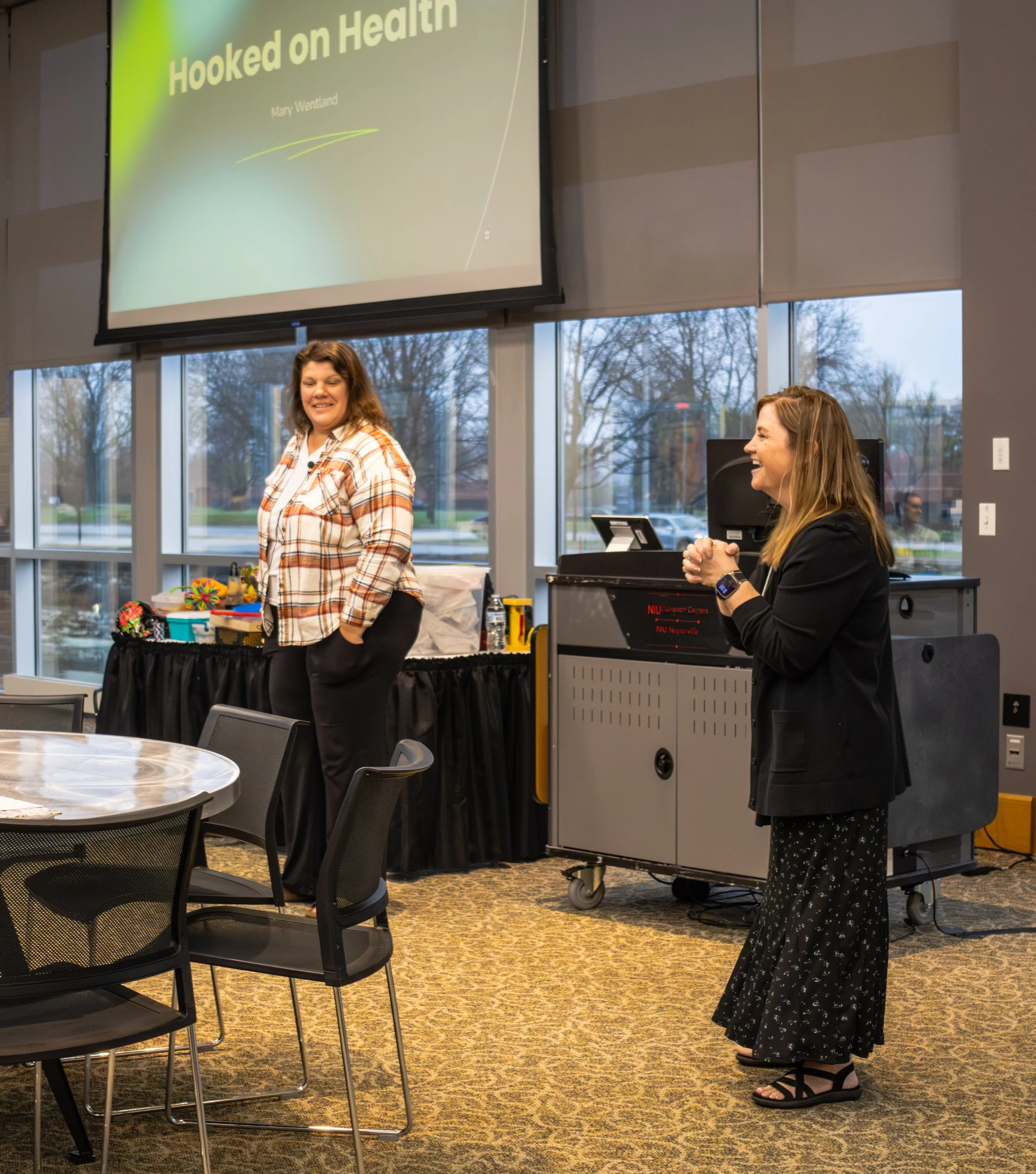 Two women standing in a conference room with large windows and a presentation screen. One woman is smiling and the other is speaking, dressed casually, surrounded by tables, chairs, and presentation equipment.