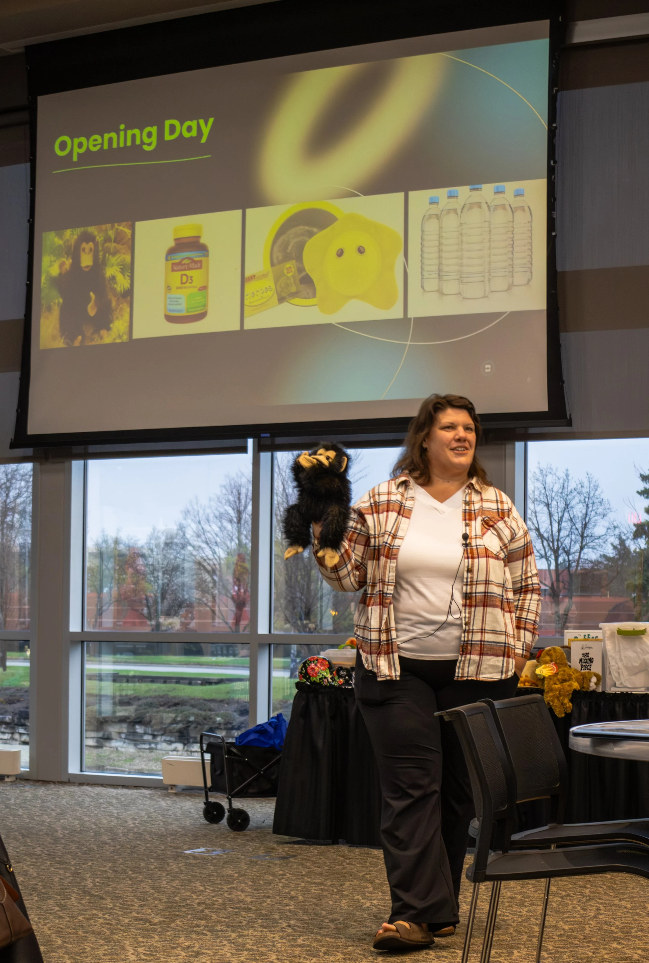 Woman standing in front of a large screen displaying a presentation slide titled 'Opening Day' with images of a bear mascot, vitamins, a yellow plush toy, and bottled water, in a room with large windows showing trees outside.