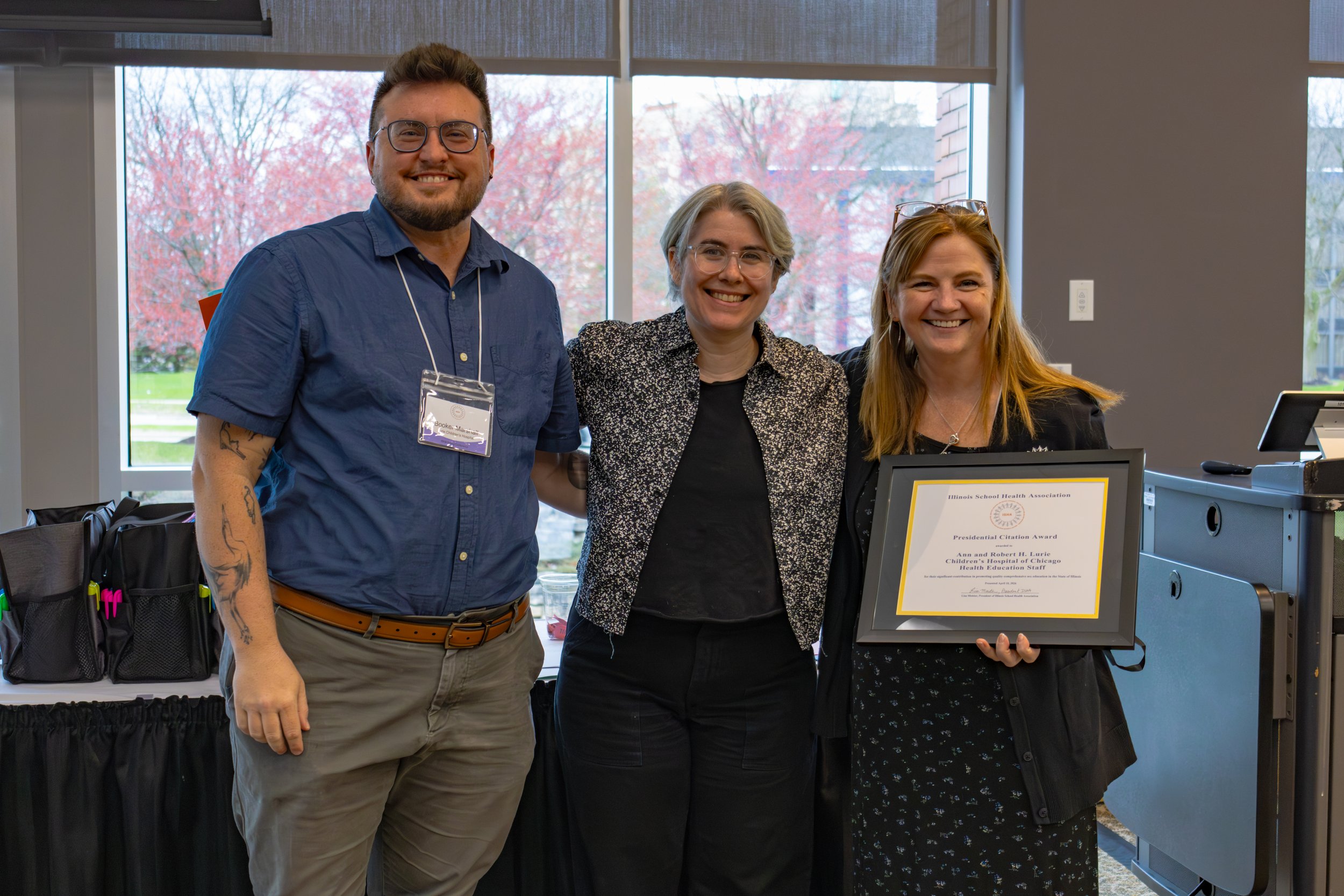 Three smiling women and a man standing together at a recognition event inside a room with large windows. One woman holds a framed award, and a certificate on a stand reads 'Illinois School Health Association.' The man wears glasses, a blue shirt, and has tattoos on his arm, while the women have light-colored hair and are dressed in professional attire.