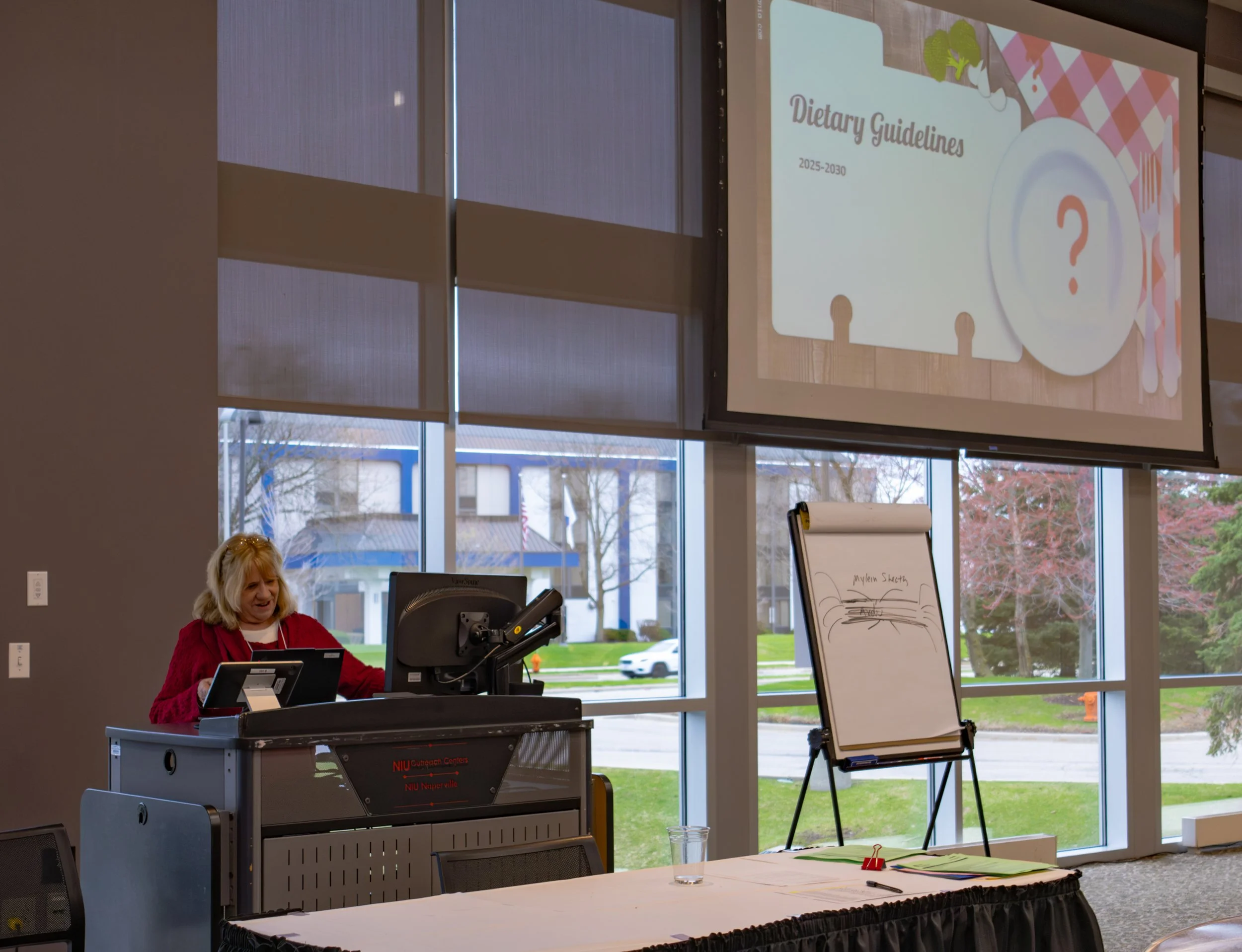 A woman in a red sweater is standing at a microphone in a room with large window, presenting slides on a large screen. The slide reads "Dietary Guidelines 2025-2030" with illustrations including a plate and a question mark. A whiteboard and papers are on a table in front of her.