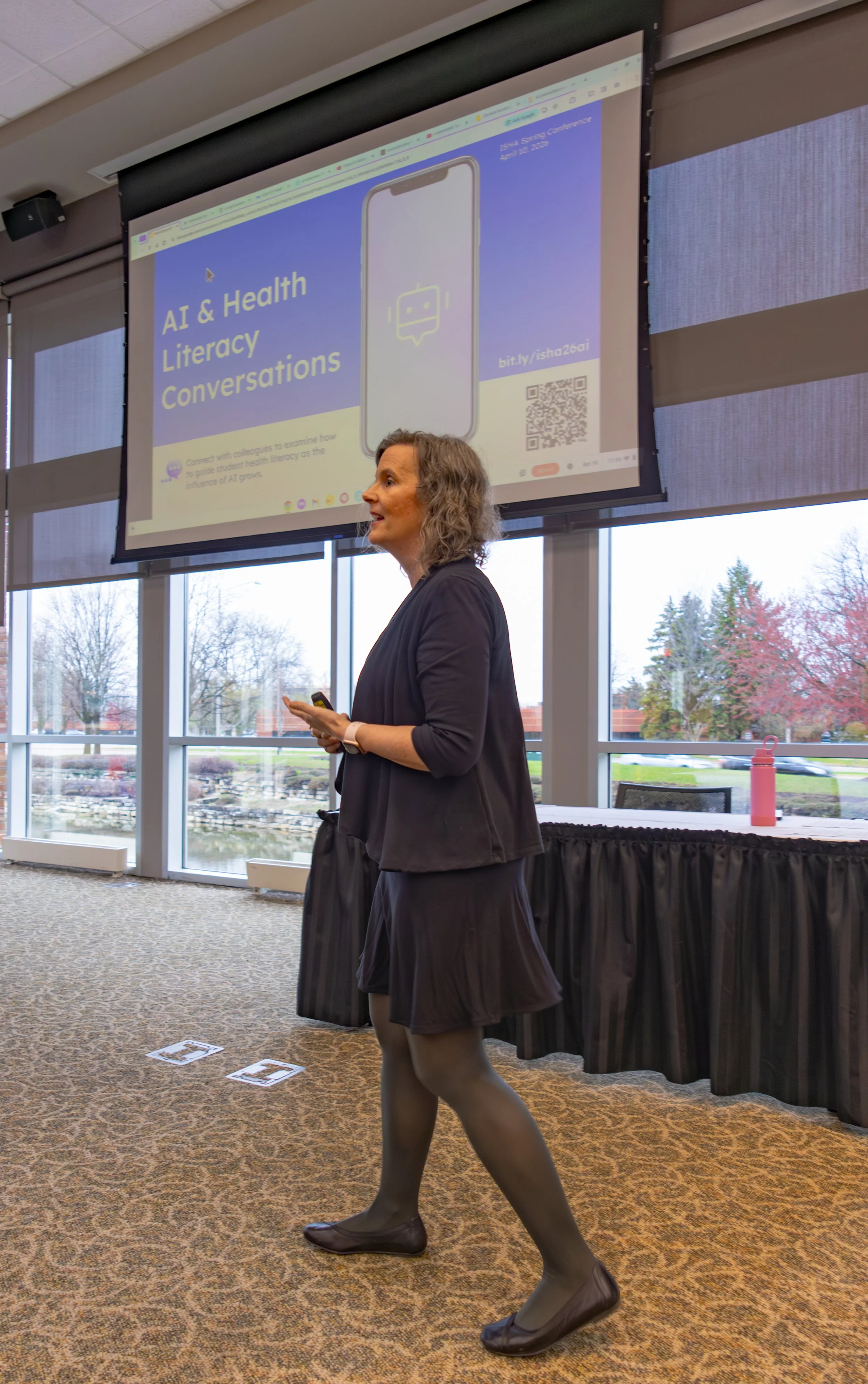 A woman in professional attire walking in front of a presentation screen at a conference. The screen displays a slide titled 'AI & Health Literacy Conversations' with an illustration of a smartphone and a QR code.