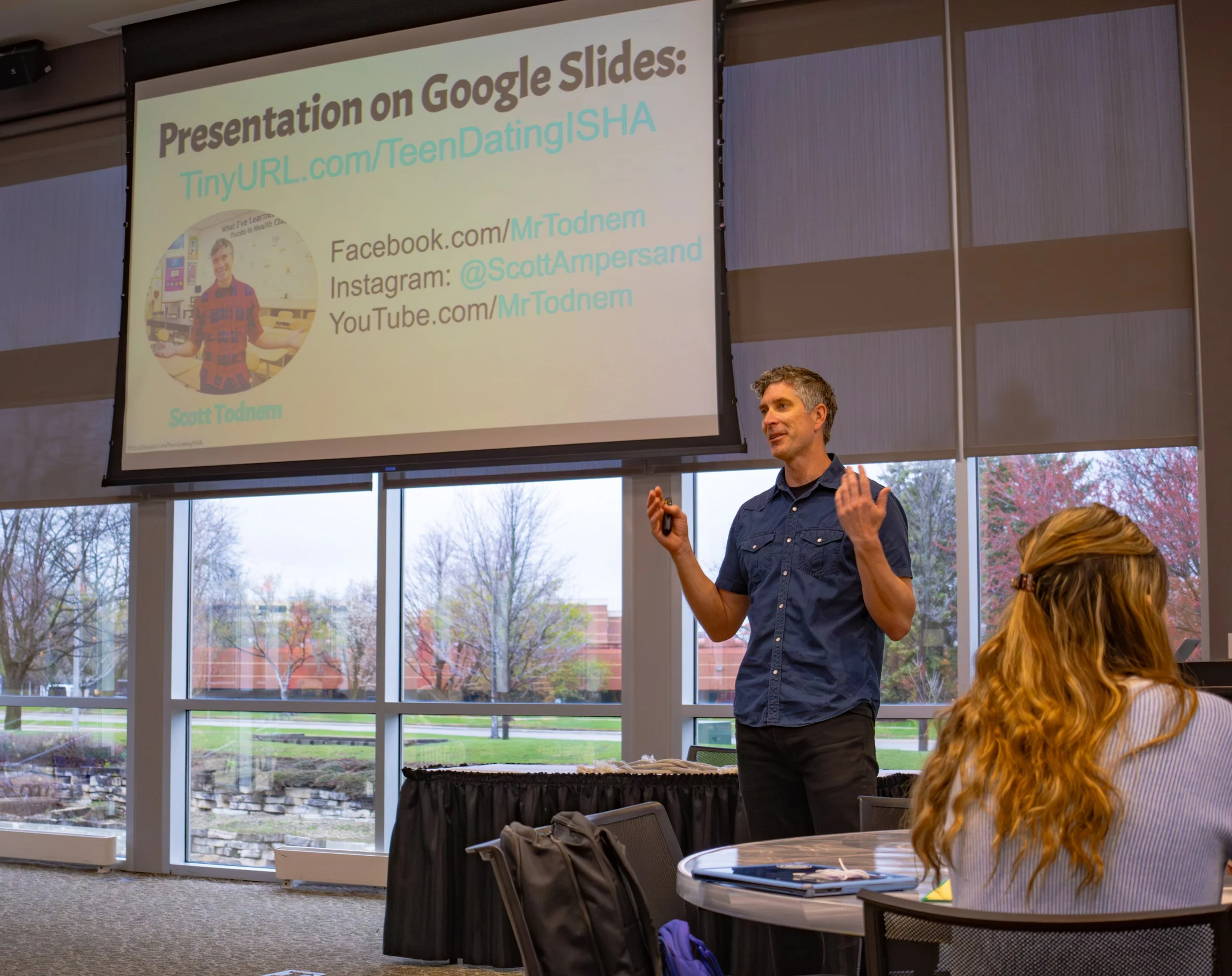 A man giving a presentation in a room with large windows, showing a slide on Google Slides with contact information. The man is wearing a dark blue shirt, and there is a woman with long hair sitting and listening. The room has a view of trees and buildings outside.