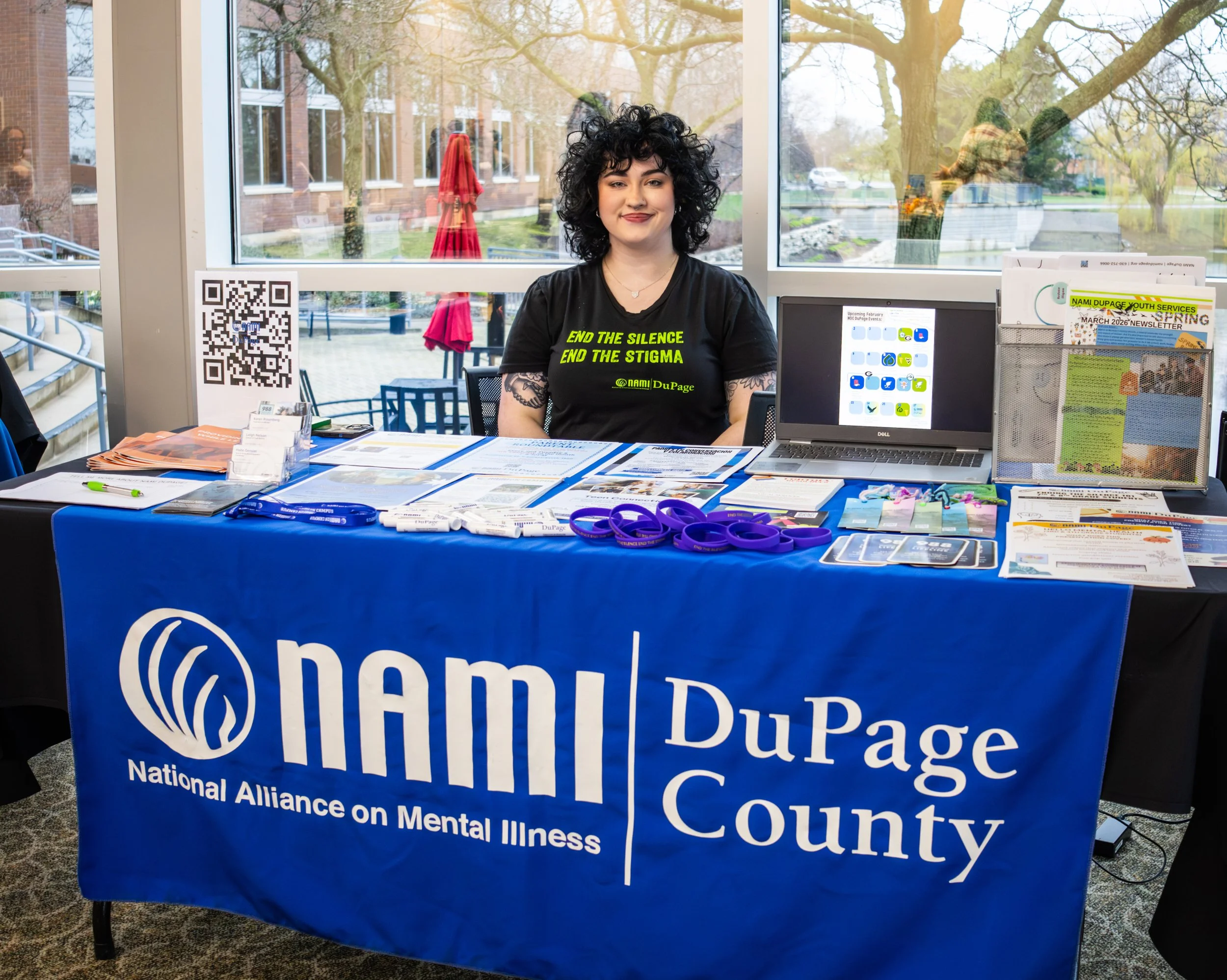 A woman sitting behind a table with a blue tablecloth displaying the words 'National Alliance on Mental Illness' and 'NAMI DuPage County'. She has short, curly black hair, wears a black T-shirt with yellow text, and is smiling. The table has various informational materials, and there is a QR code on the left side. Behind her, there are large windows showing trees and a building outside.