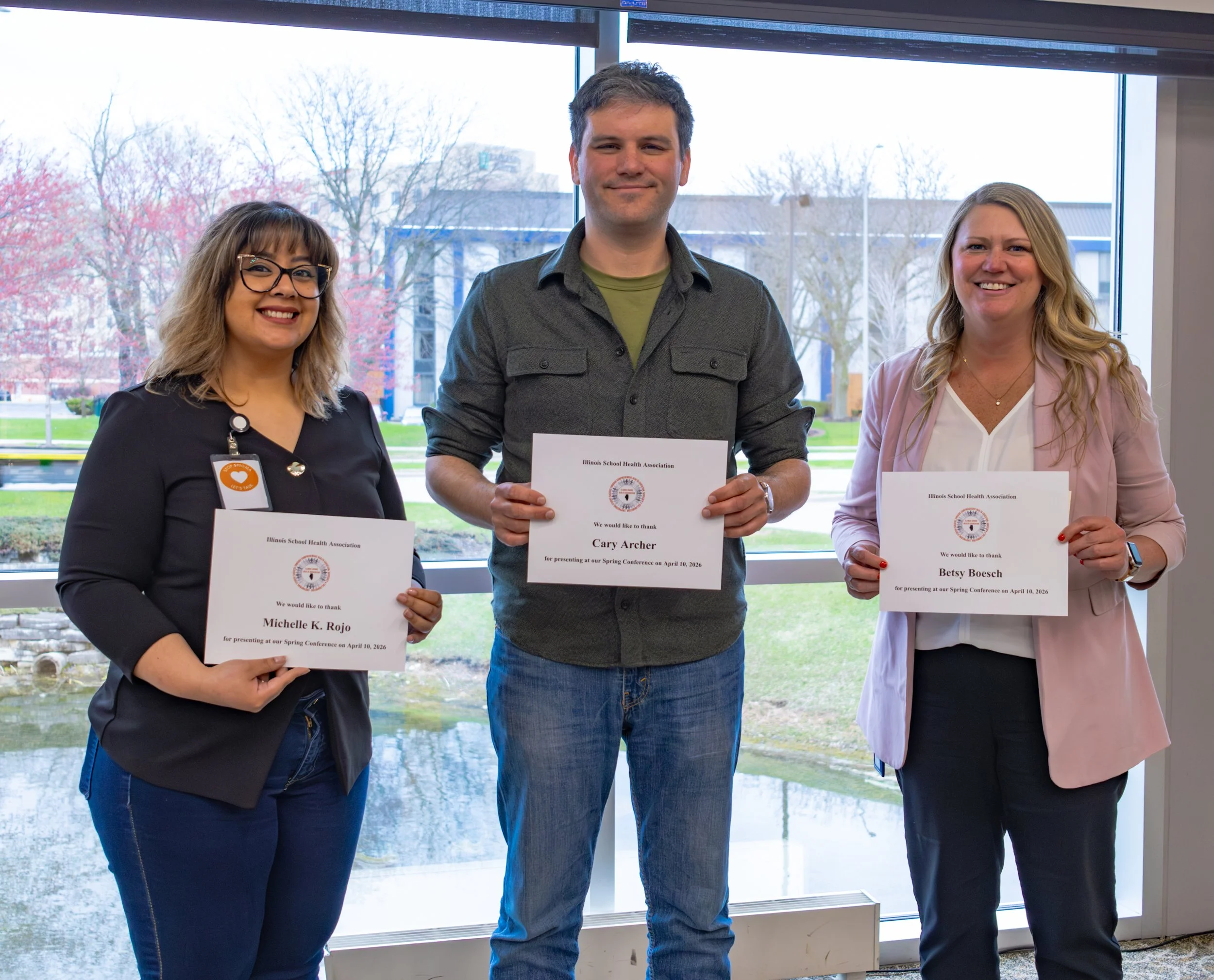 Three individuals standing indoors in front of a large window, holding certificates of appreciation from the Illinois School Health Association, smiling for a group photo during a formal event.