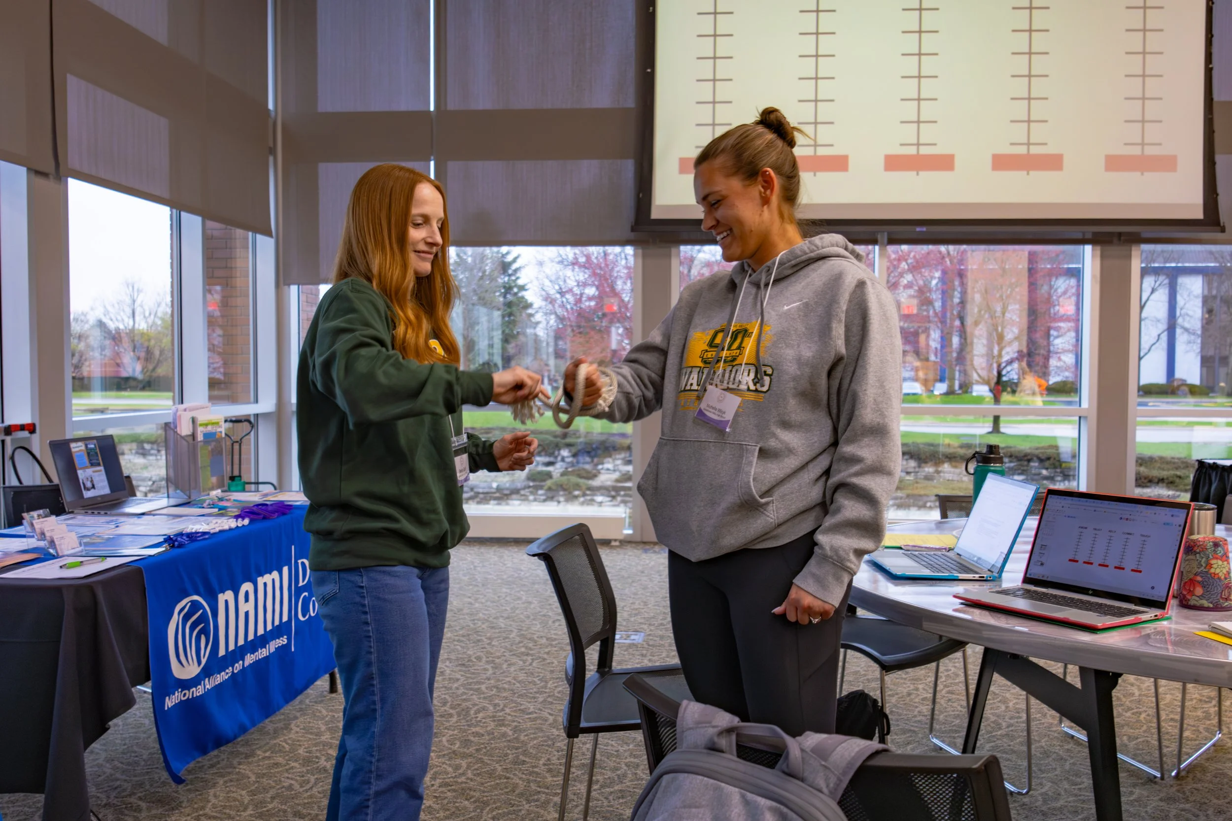 Two women exchanging a handshake at a conference or event, with a table displaying promotional materials and a large screen in the background showing tall charts or graphs.