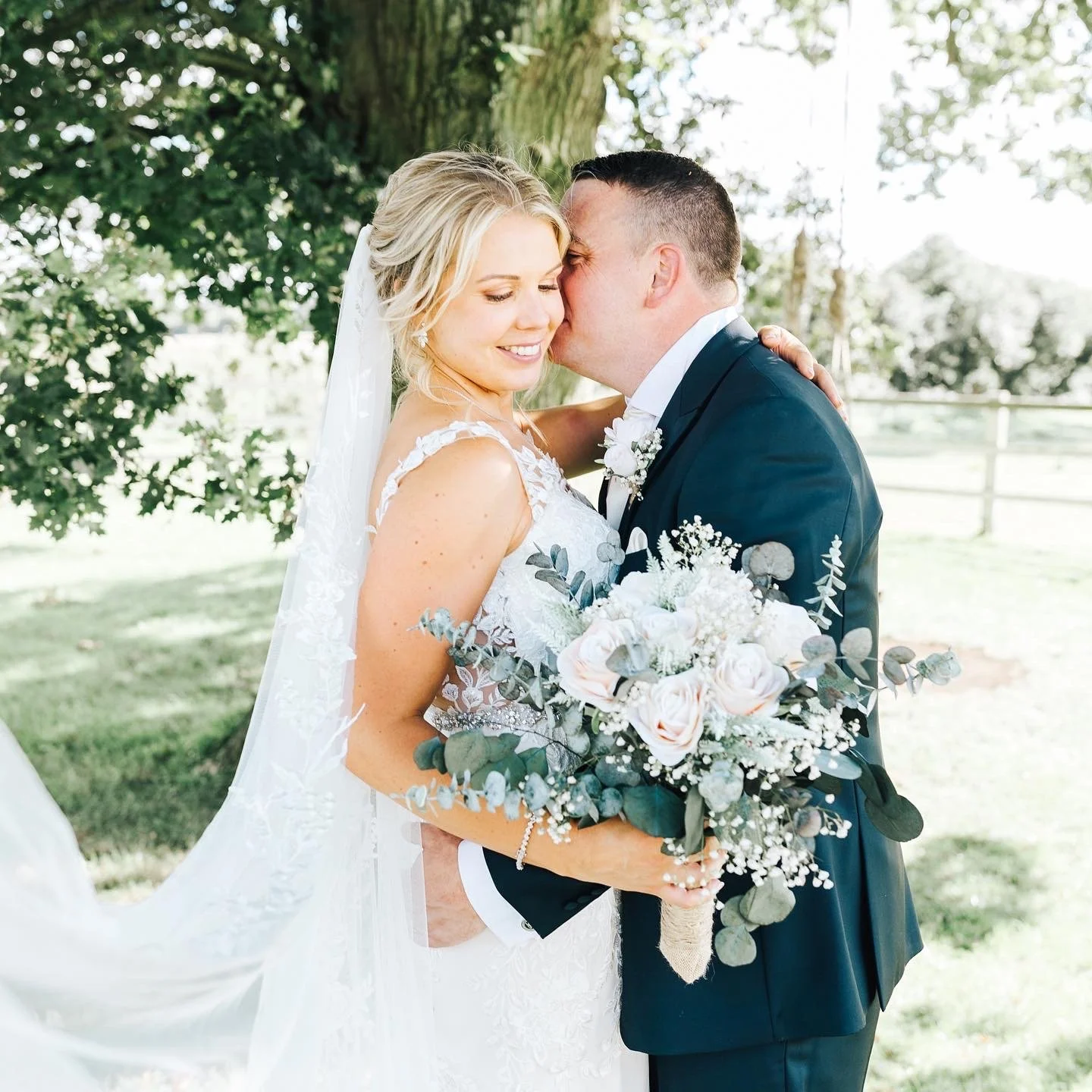 Bride and groom embracing outdoors, with the groom whispering into the bride's ear, the bride holding a bouquet of white and pale pink roses and greenery, under a tree with a fence in the background.