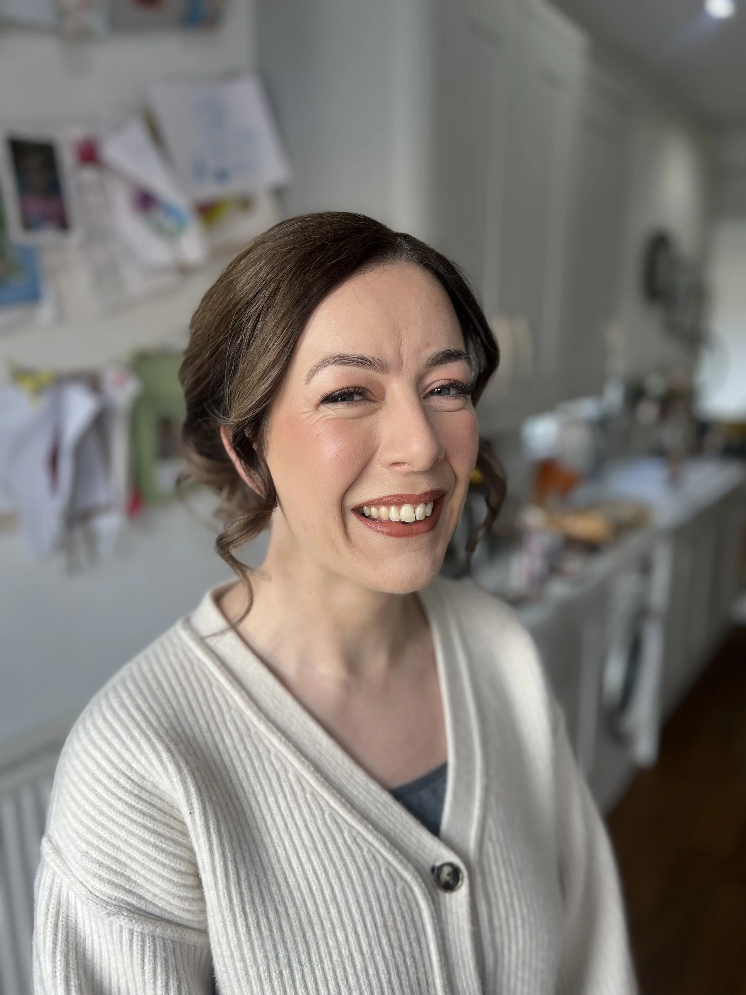 A woman with brown hair styled in loose curls, smiling and winking, wearing a light-colored cardigan over a top, standing in a kitchen with a blurred background.