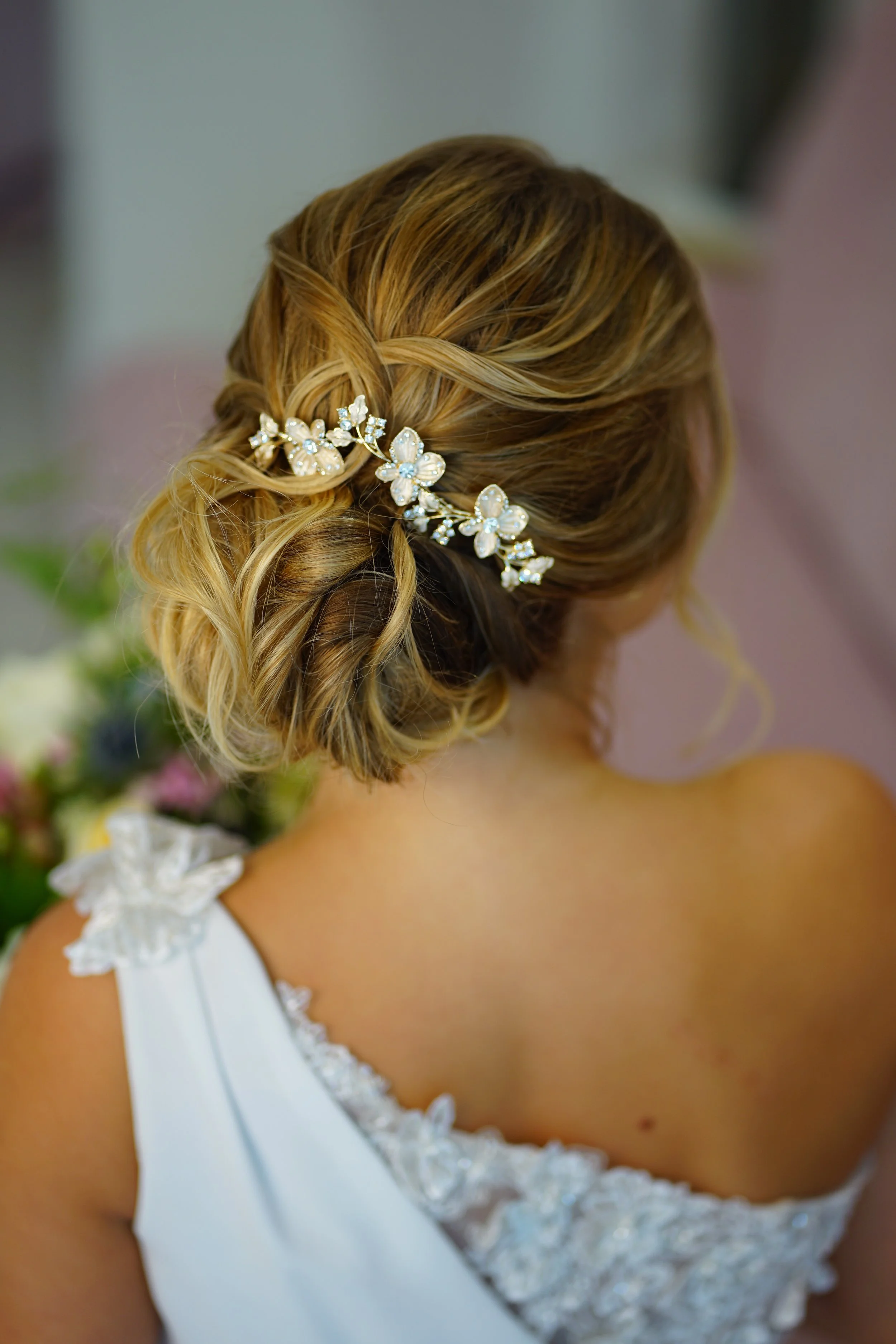 Bride with styled blonde hair in loose curls, adorned with a floral hairpiece, wearing a white gown with lace details on the shoulder.