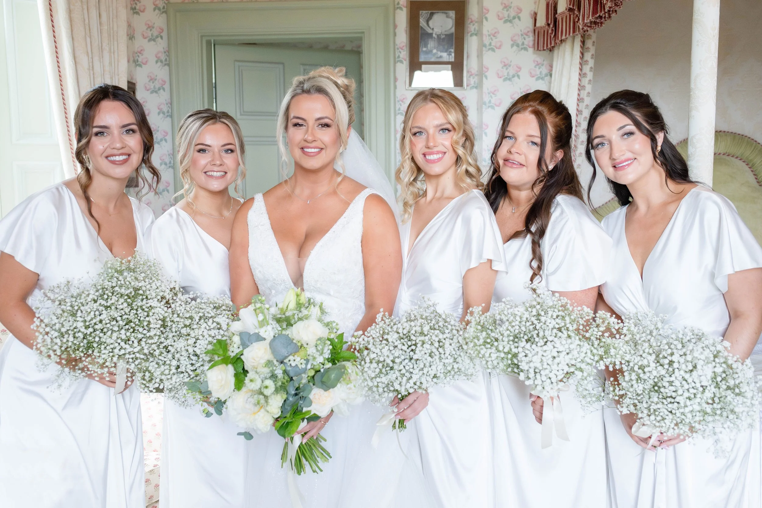 Seven women dressed in white gowns, likely at a wedding, standing indoors in front of floral wallpaper and curtains, with the central woman holding a bouquet of white and green flowers.