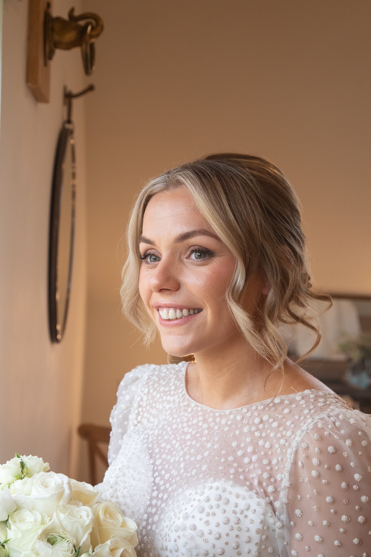 A smiling woman with blonde hair styled in loose waves, wearing a white dress with textured polka dot pattern, holding a bouquet of white roses, in a warmly lit room with a neutral background.