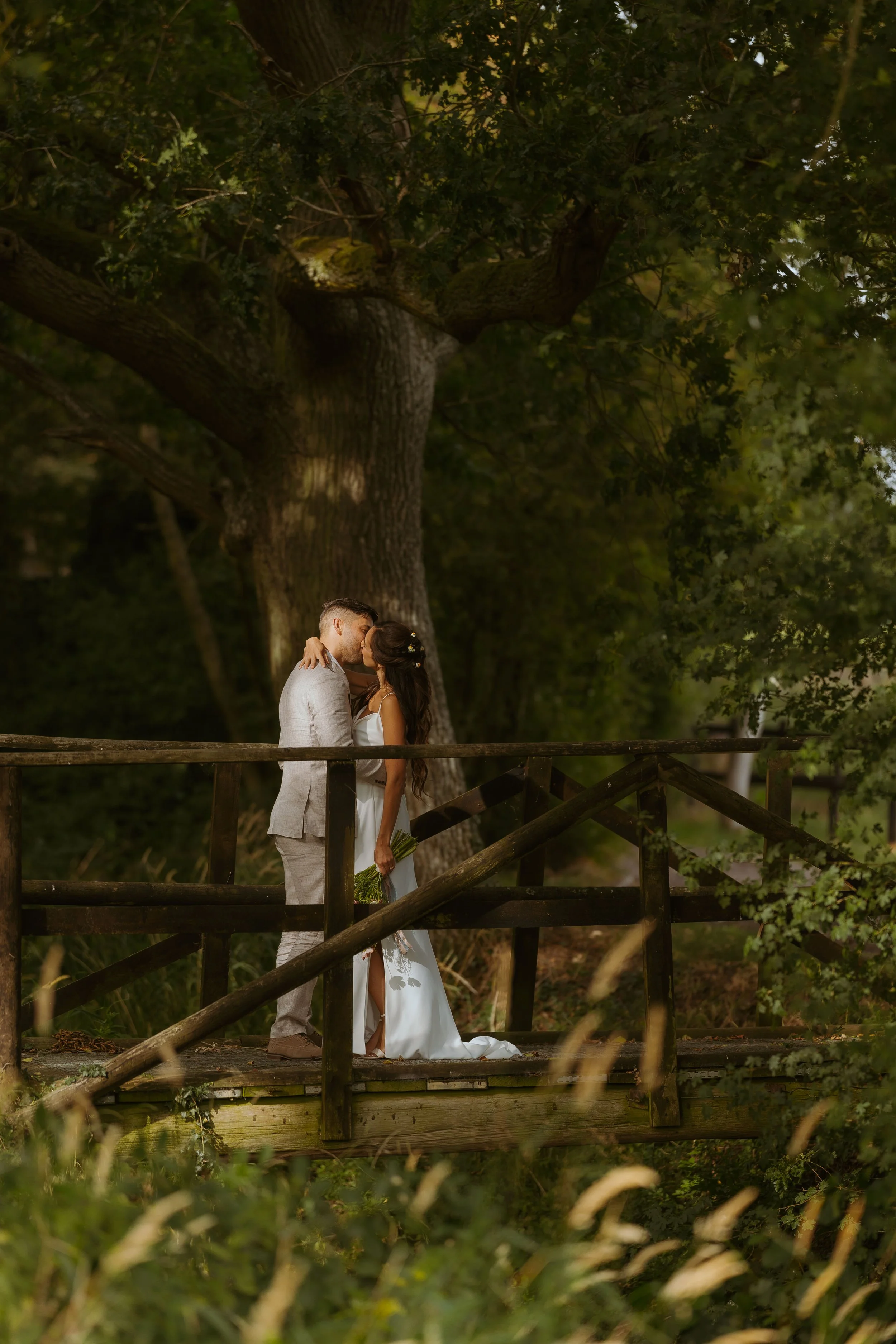 A couple dressed in wedding attire sharing a kiss on a wooden bridge surrounded by lush green trees.