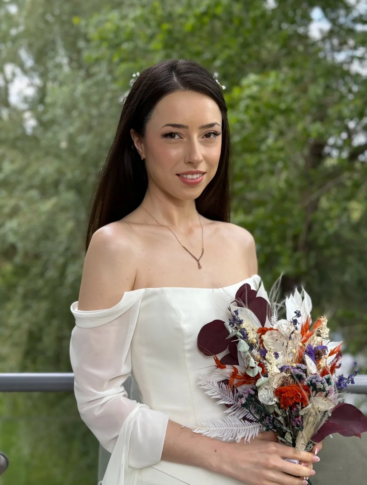 A woman in a white off-shoulder dress holding a colorful bouquet of flowers outdoors with green trees in the background.