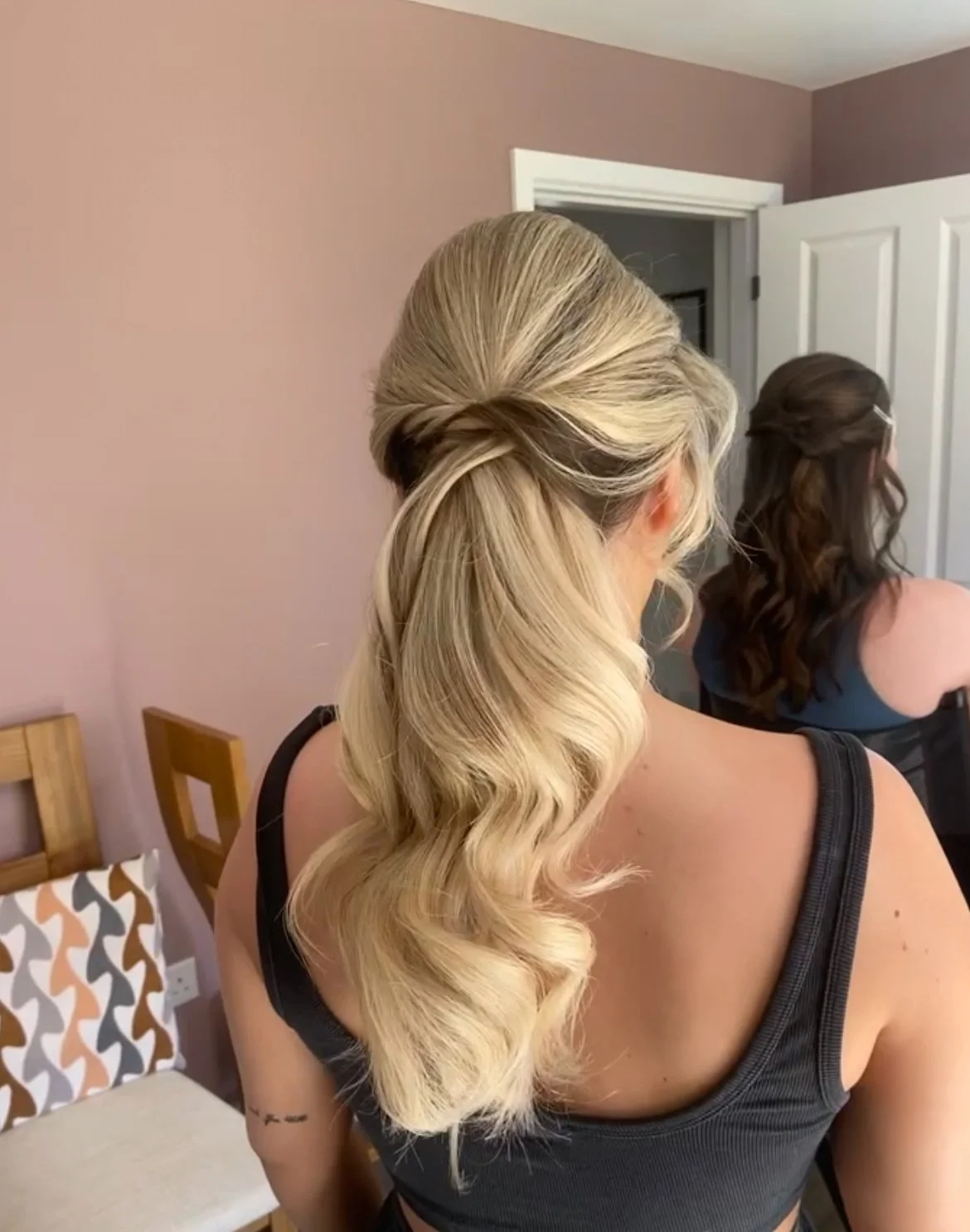 A woman with styled blonde hair, curled in soft waves, seen from the back in a bedroom.