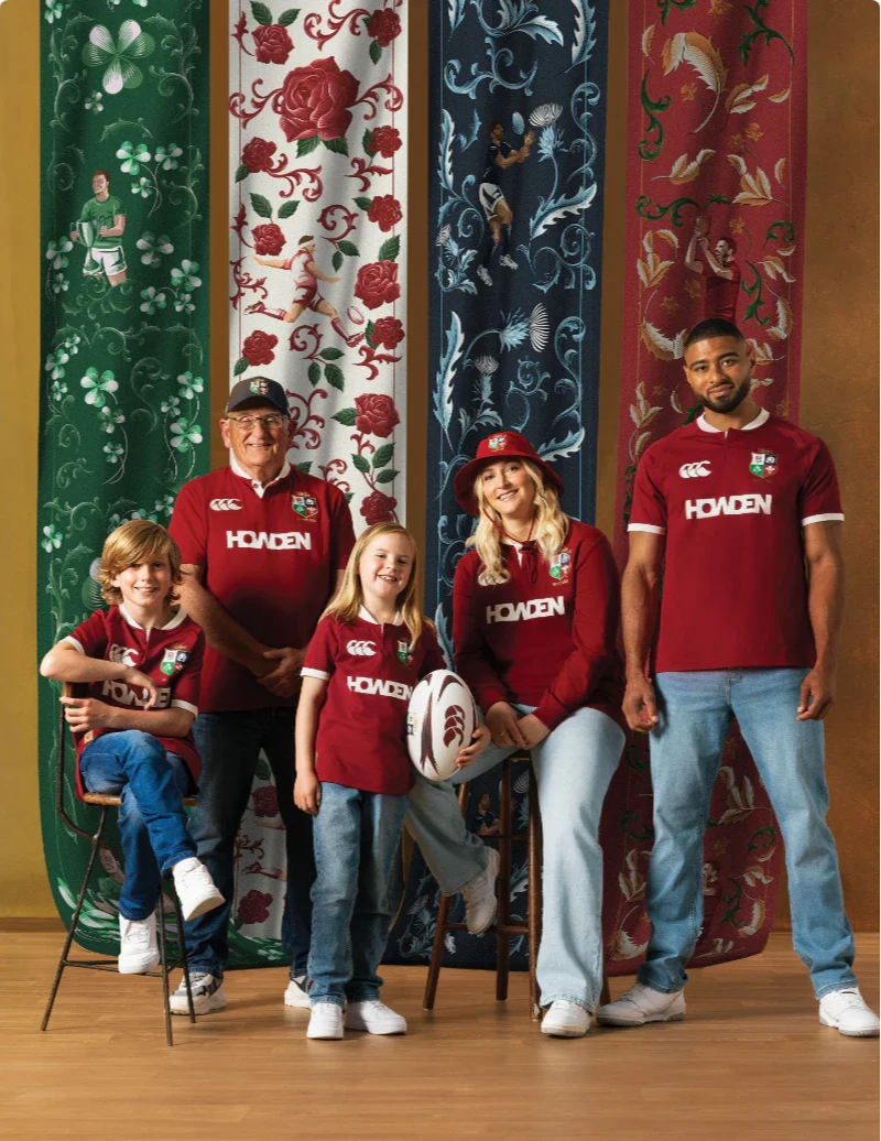 Group of five people, including two children, an older man, a young woman, and a young man, wearing matching red sports jerseys with 'HODEN' and a logo, standing and sitting in front of decorative panels with floral and figure designs, indoors on a w