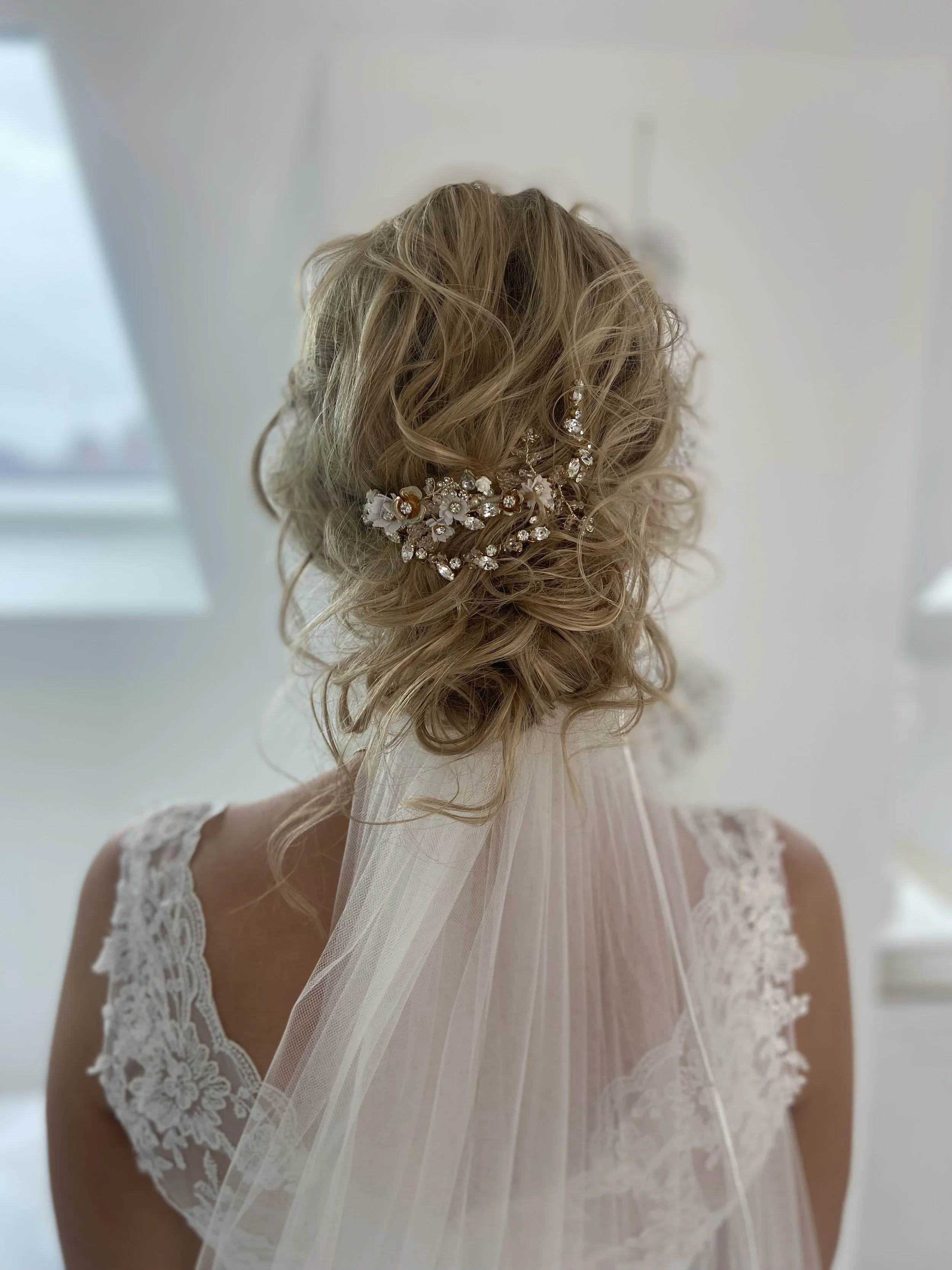 A woman with styled, curly blonde hair wearing a lace wedding dress with a sheer veil and a floral hair accessory with rhinestones.