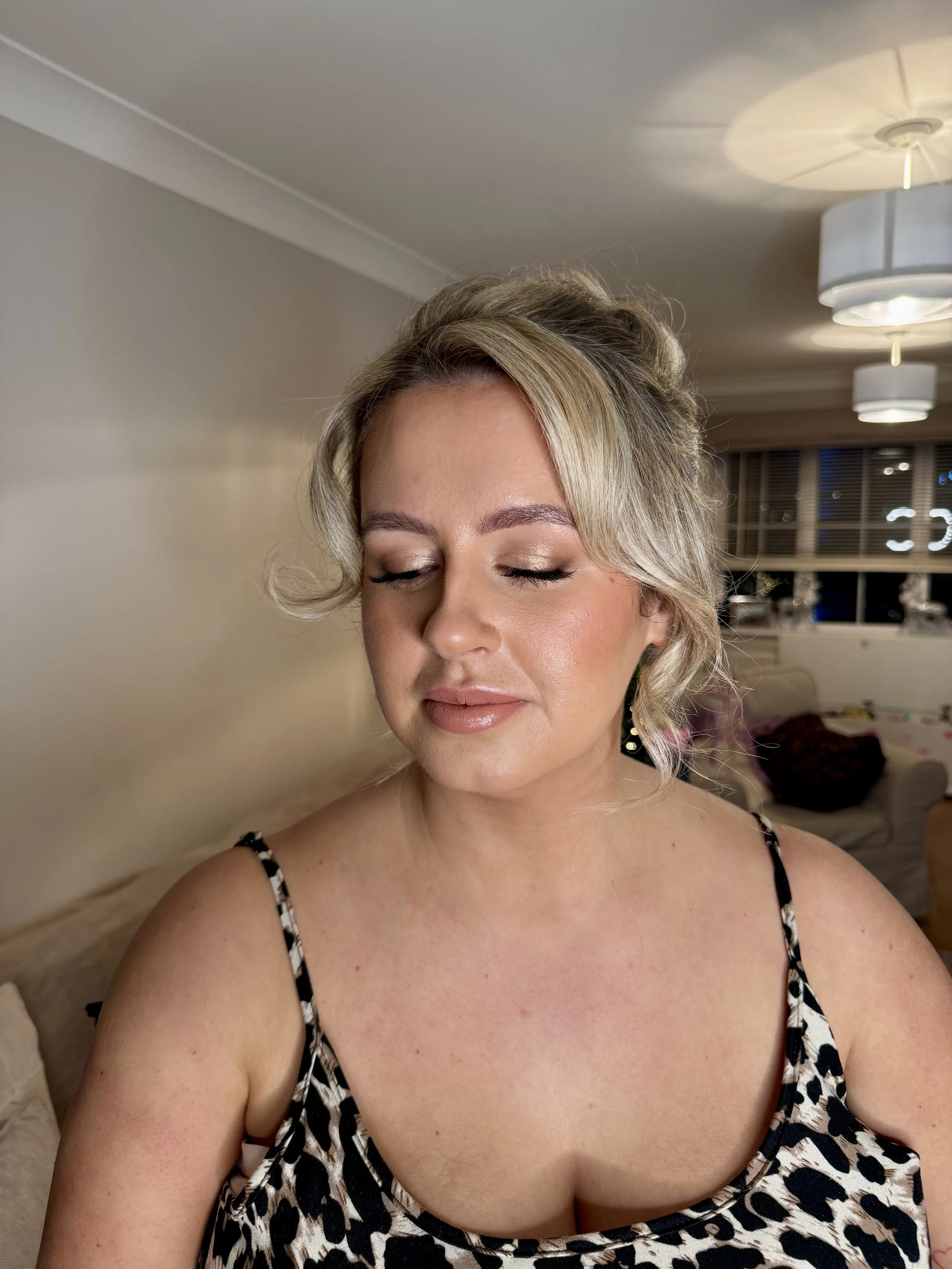 A woman with blonde hair styled in loose waves, wearing makeup, a black and white leopard print dress, and gold earrings, with closed eyes, in a warmly lit living room.