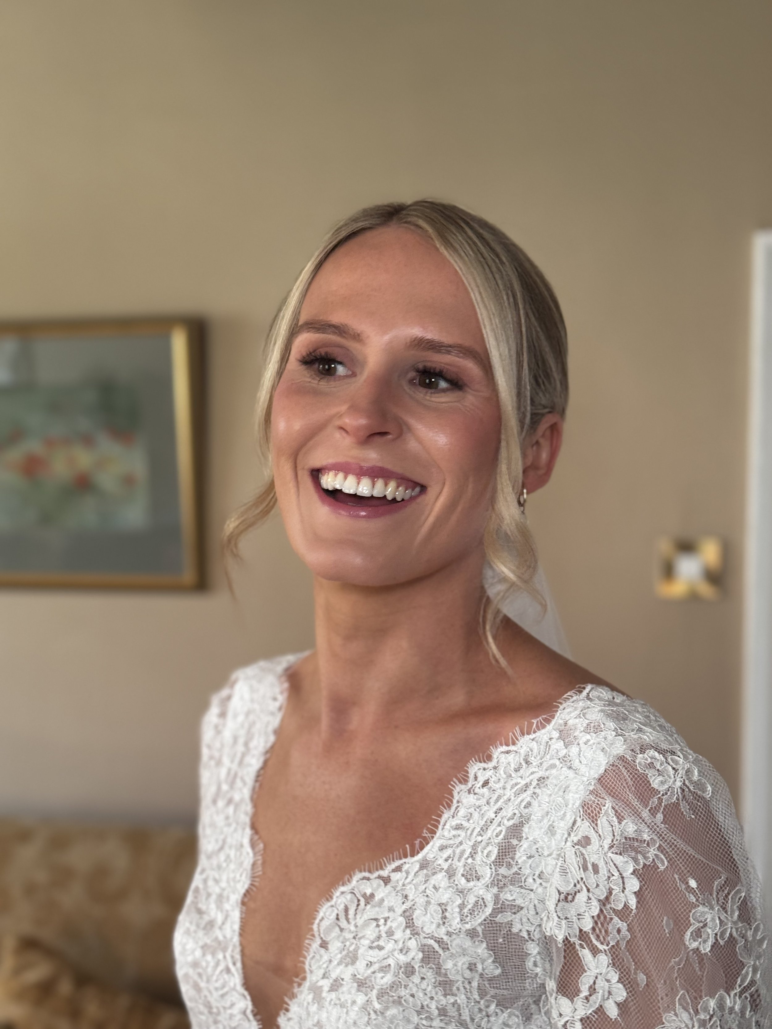 A smiling woman in a lace wedding dress with blonde hair styled in a loose updo, standing indoors with a framed artwork in the background.