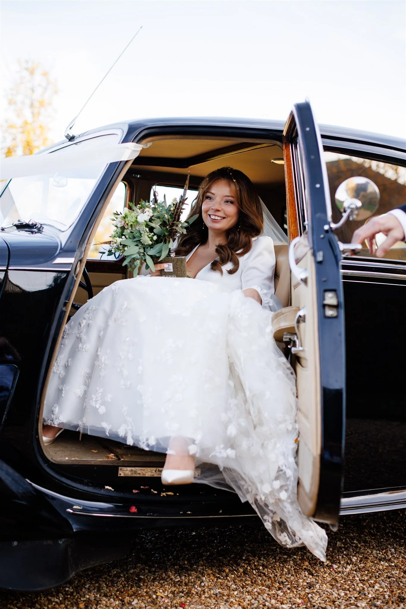 Bride sitting in a vintage black car, holding a bouquet, smiling, wearing a white wedding dress with floral embroidery, and with the car door open.