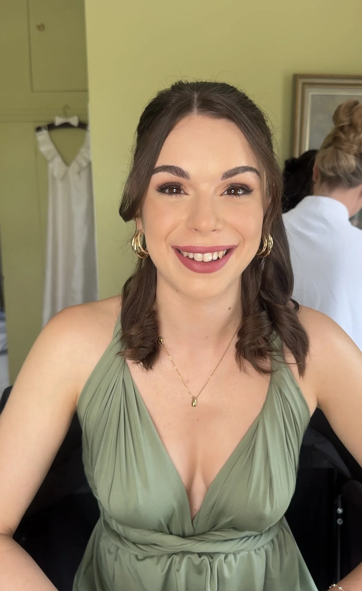 Close-up of a woman smiling, wearing a green sleeveless dress, gold jewelry, and sitting indoors.