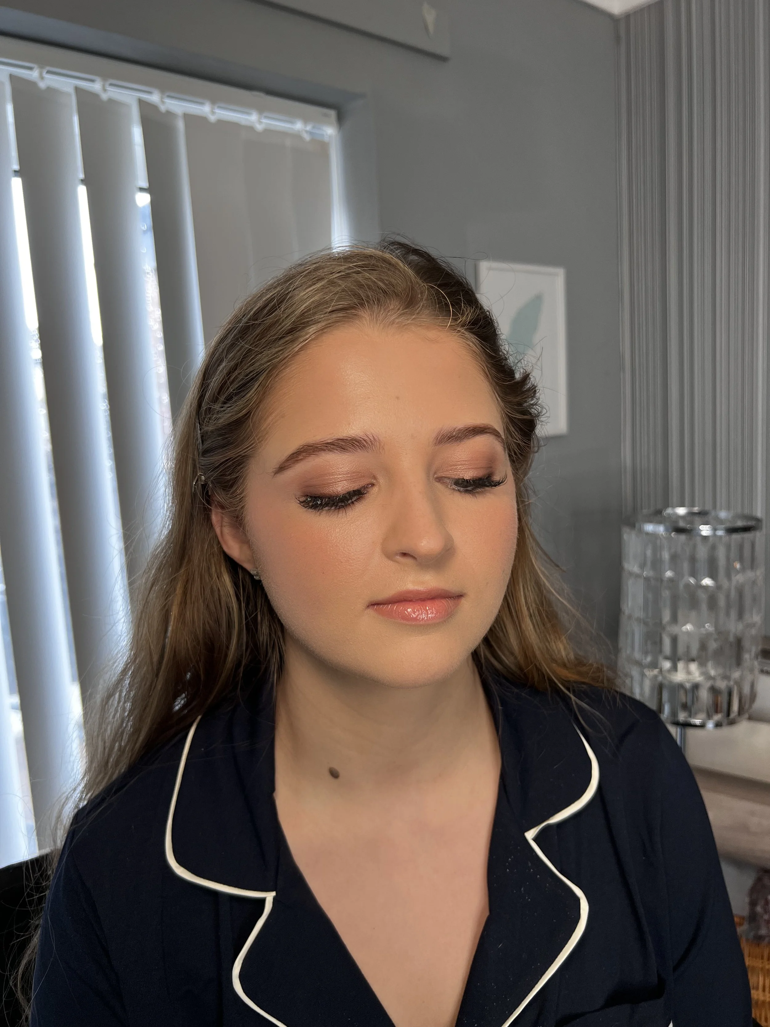 A young woman with closed eyes and flawless makeup, wearing a navy blue shirt with white piping, is seen in a room with vertical blinds and a framed picture on the wall behind her.