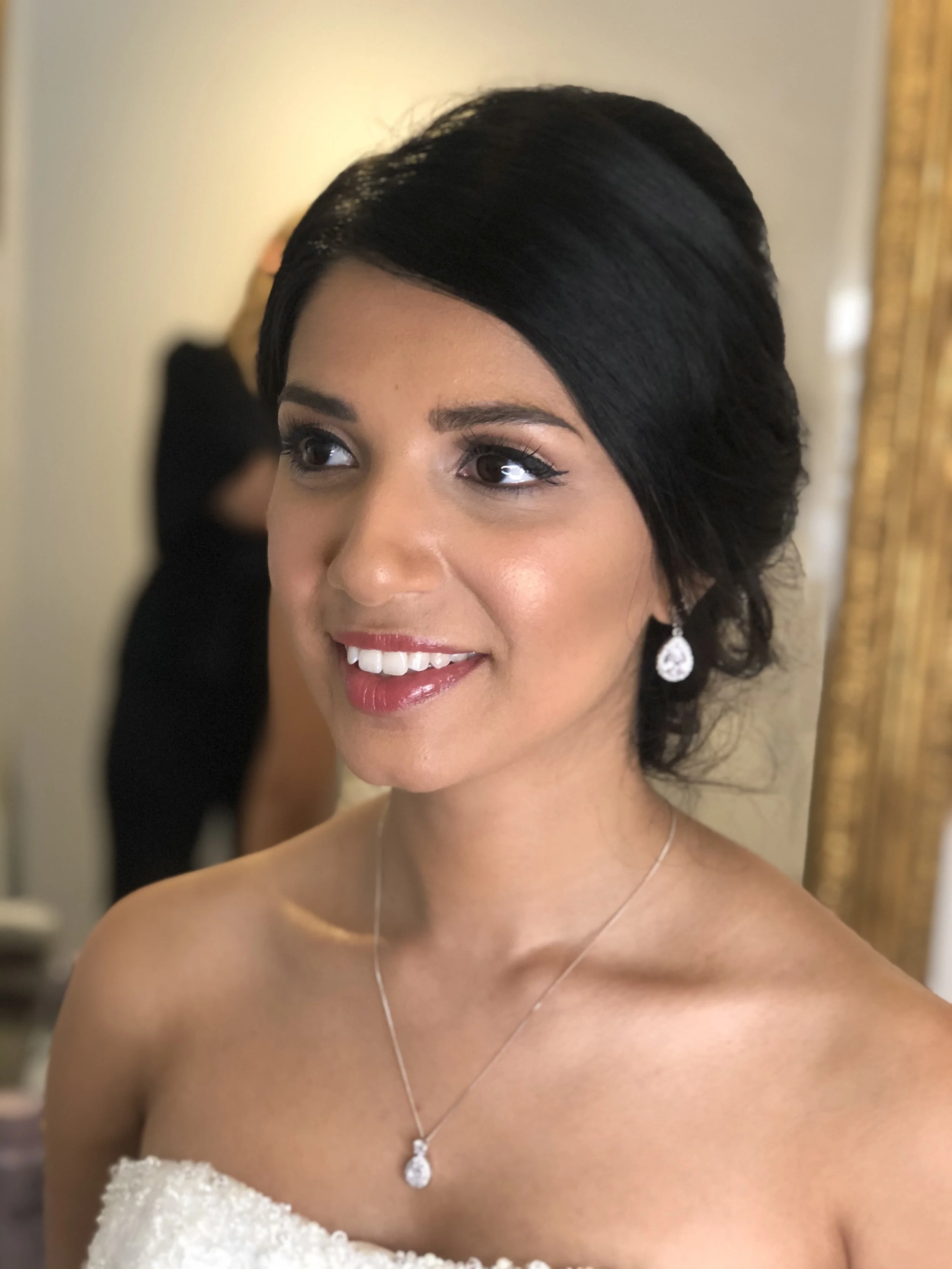 A woman with black hair styled in an elegant updo, wearing a strapless white dress with beaded details, diamond earrings, and a matching necklace, smiling and looking slightly to the side.