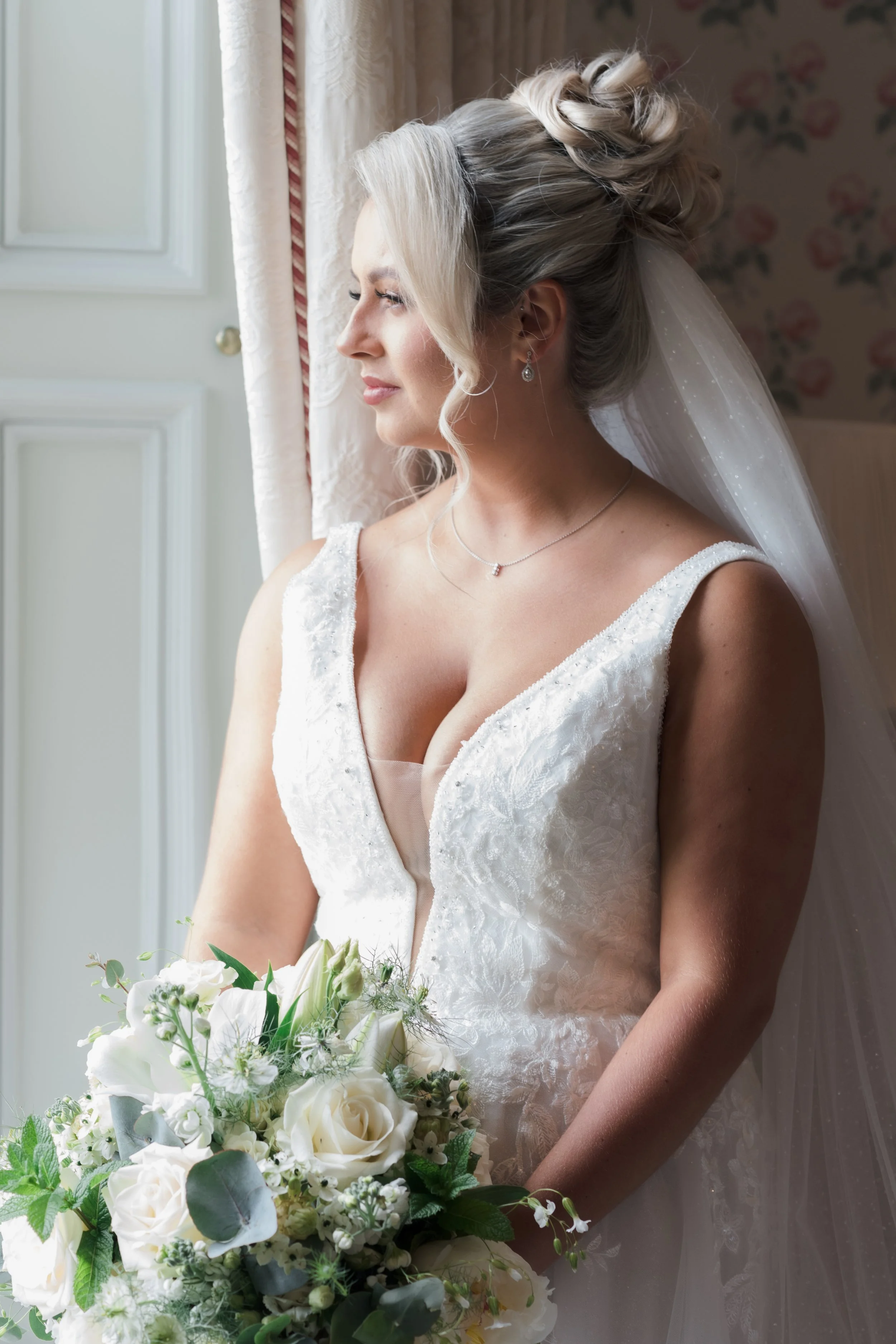A bride with platinum blonde hair styled in an elegant updo, wearing a white lace wedding dress with a deep V neckline, standing by a window with a bouquet of white roses and greenery, looking out contemplatively.