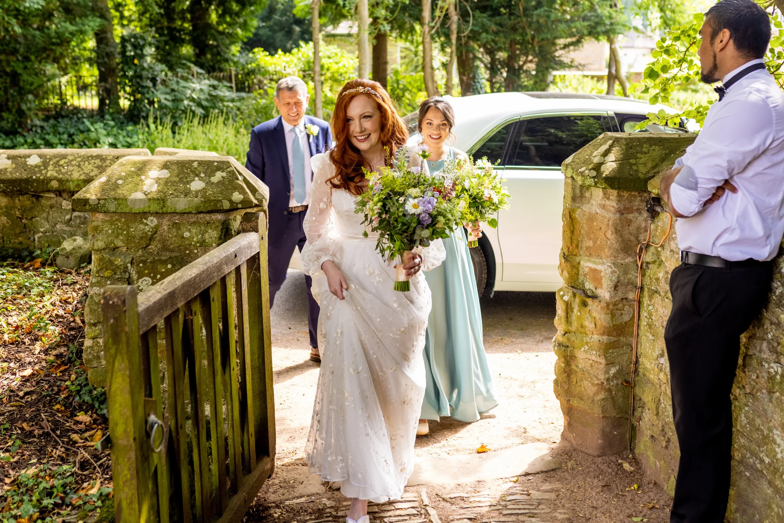 A bride with red hair in a white wedding dress holding a bouquet of flowers, walking outdoors through a garden gate, with a woman in a light blue dress and an older man in a suit behind her, and a man in a white shirt and black bow tie leaning agains