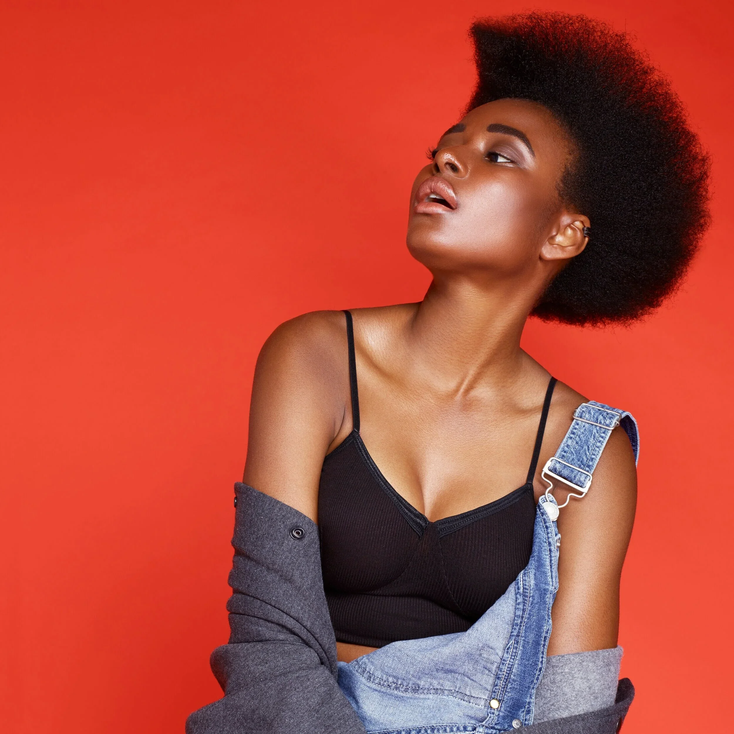 A woman with natural curly hair wearing a black top and denim overalls against an orange background.