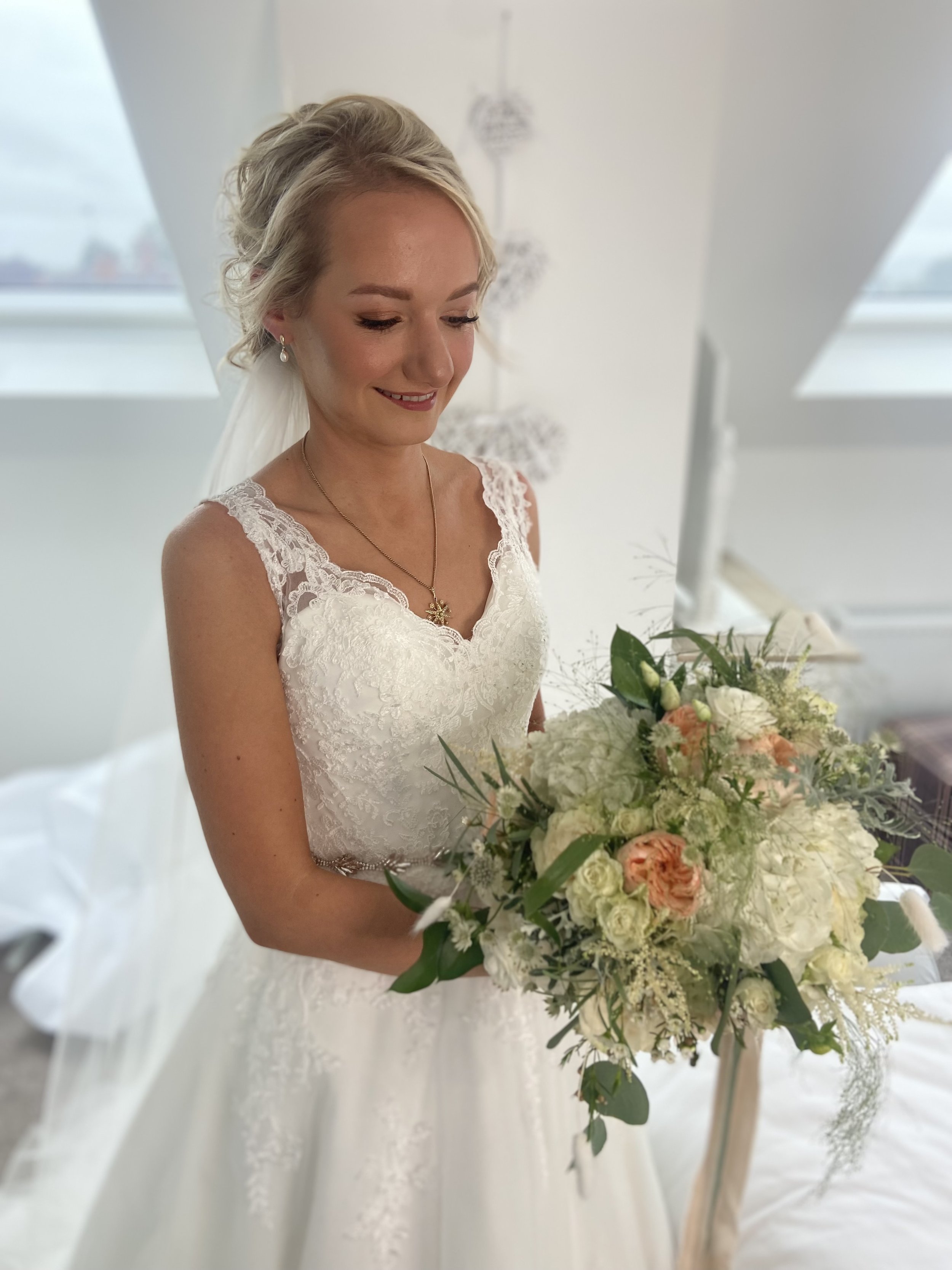A bride in a white lace wedding dress holding a bouquet of white and peach flowers, smiling and looking down.