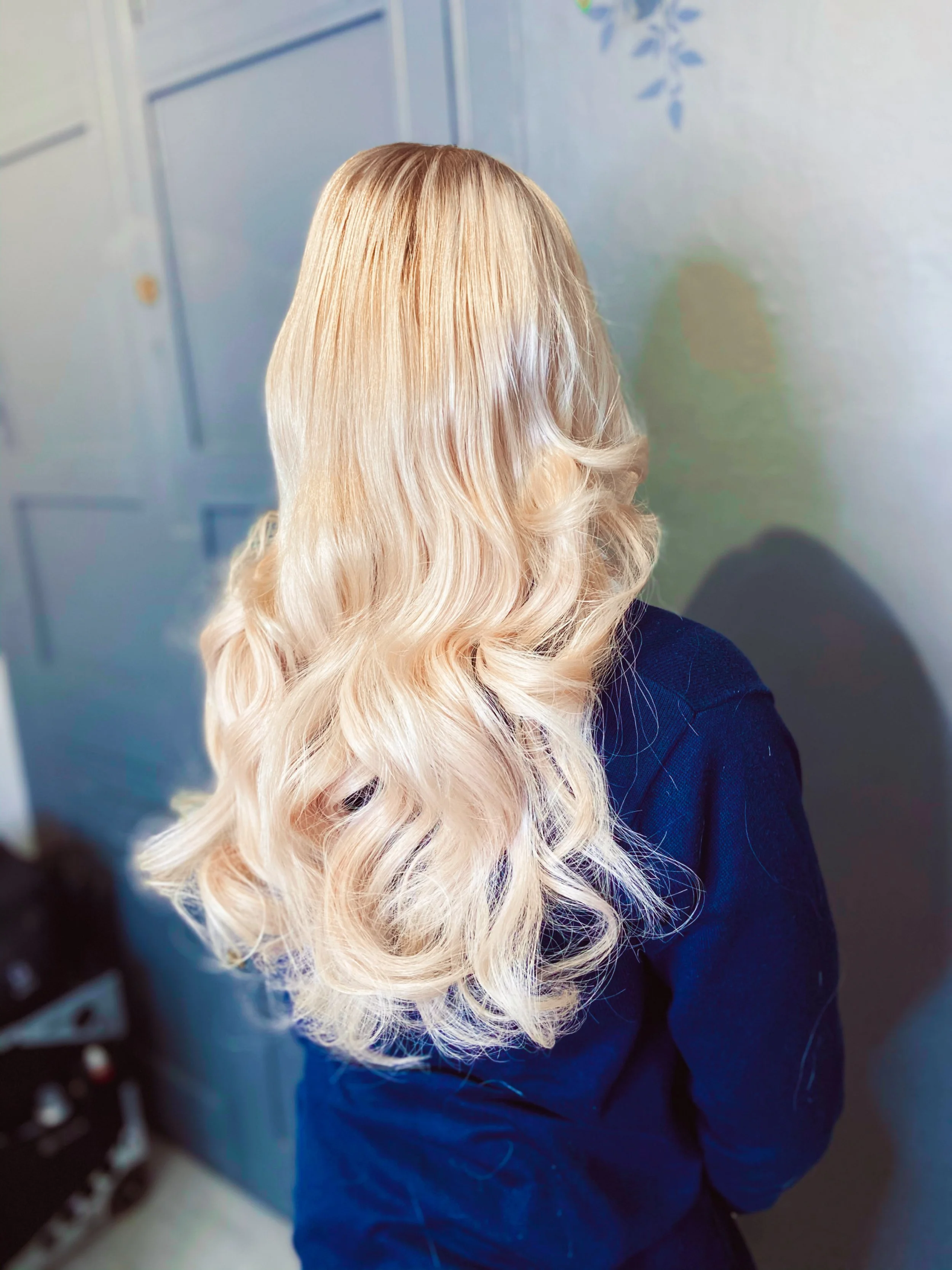 Back view of a person with long, wavy blonde hair wearing a dark blue shirt, standing indoors near a light-colored wall and door.