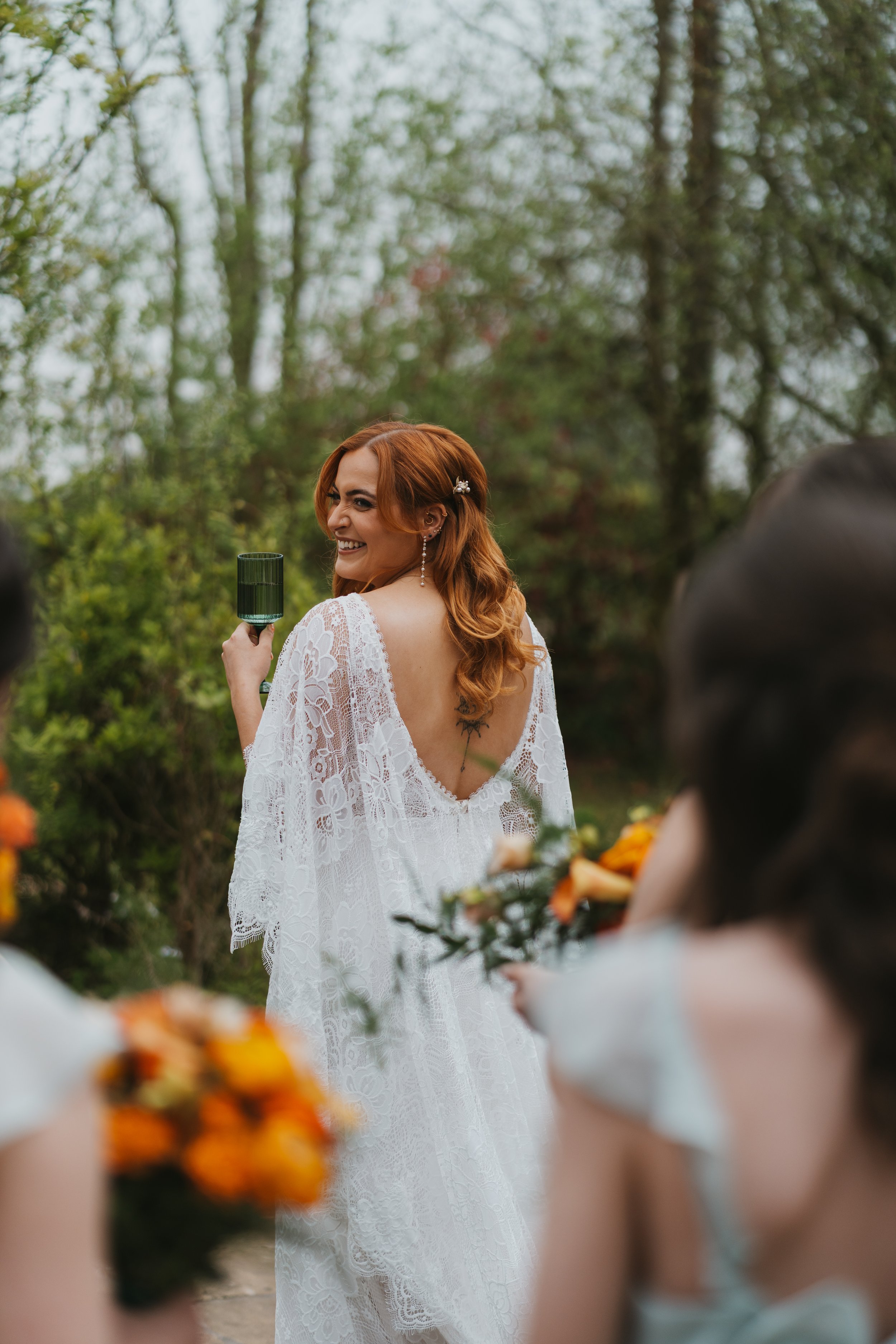 A red-haired woman in a white lace dress with a deep V back, holding a green goblet, smiling at an outdoor wedding ceremony with blurred guests holding flowers around her.