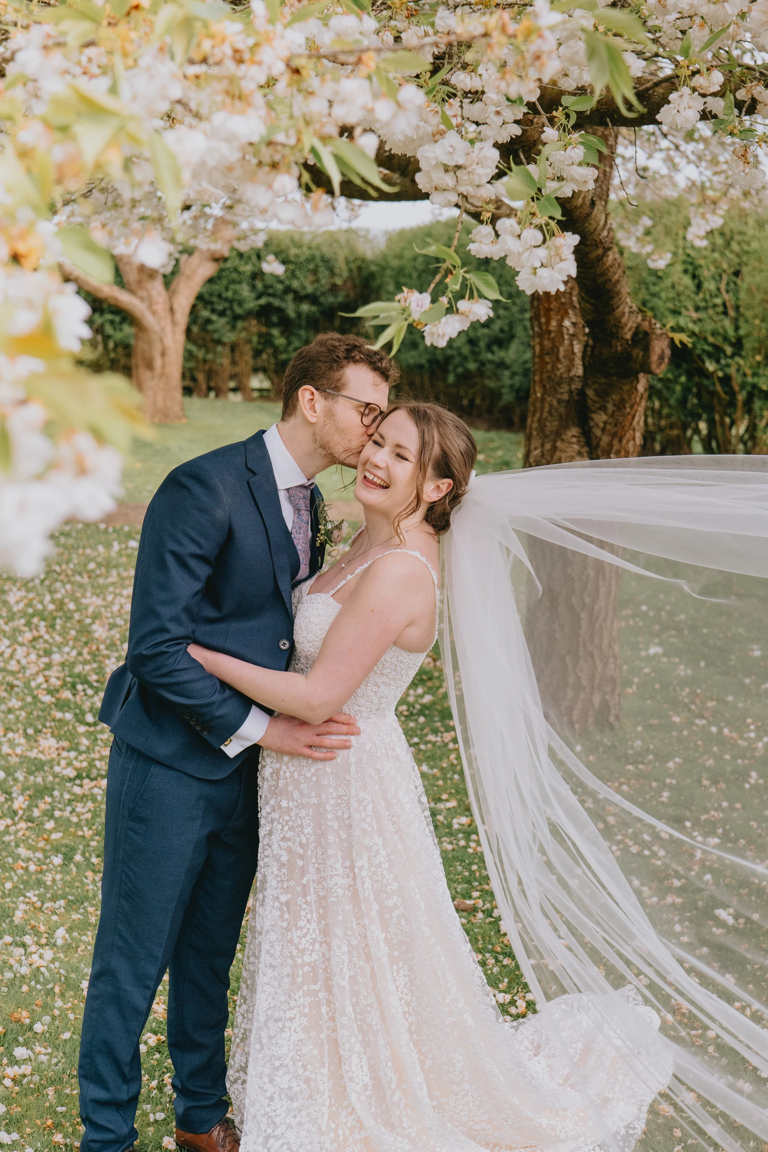 A newlywed couple standing under blossoming trees on their wedding day, sharing a joyful moment with the groom kissing the bride on the forehead.