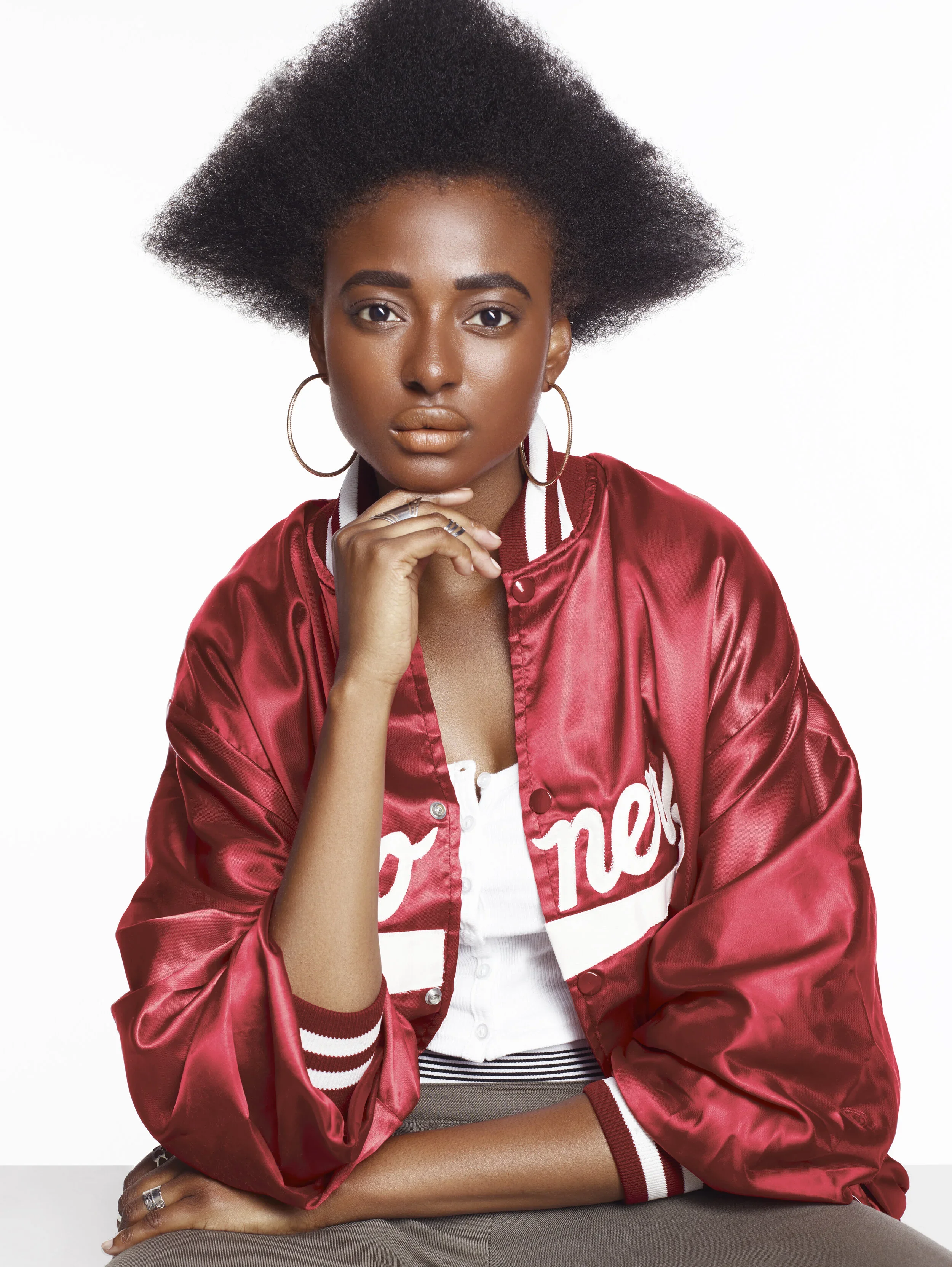 A young woman with natural hair wearing a red satin varsity jacket and hoop earrings, sitting with her hand under her chin against a plain white background.