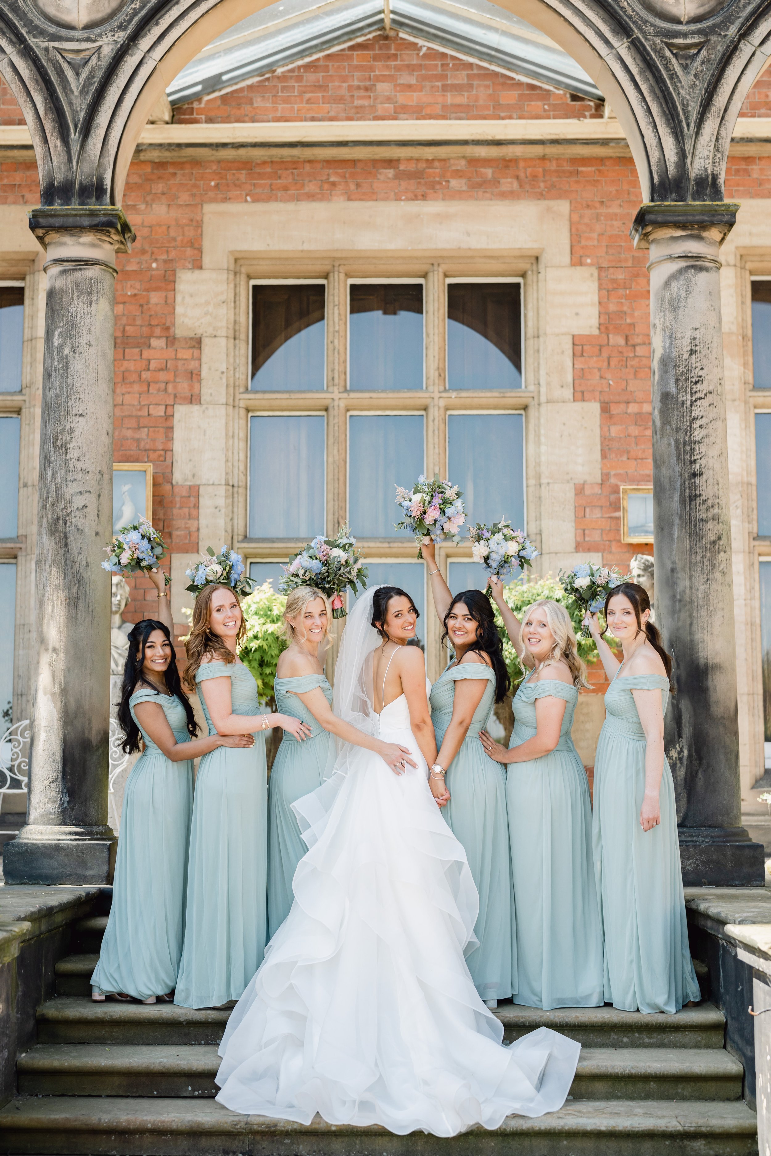Bride with six bridesmaids holding bouquets together outside a historic brick building with columns.