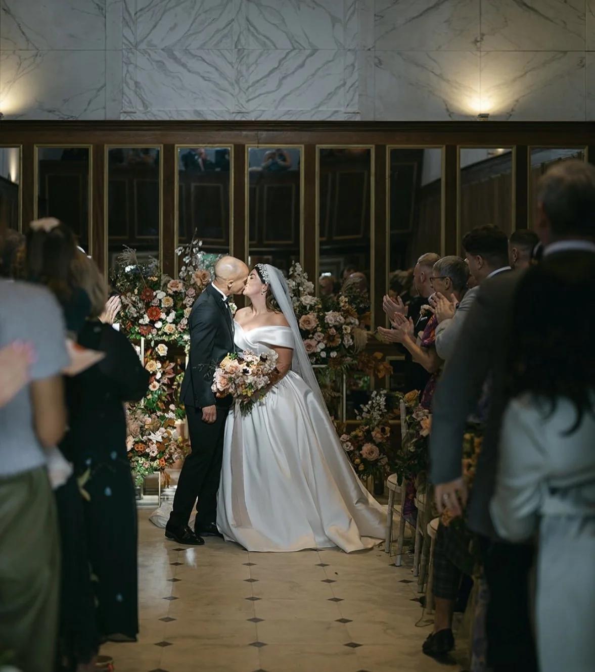 A bride and groom share a kiss during their wedding ceremony, surrounded by guests and floral decorations in an elegant indoor setting.