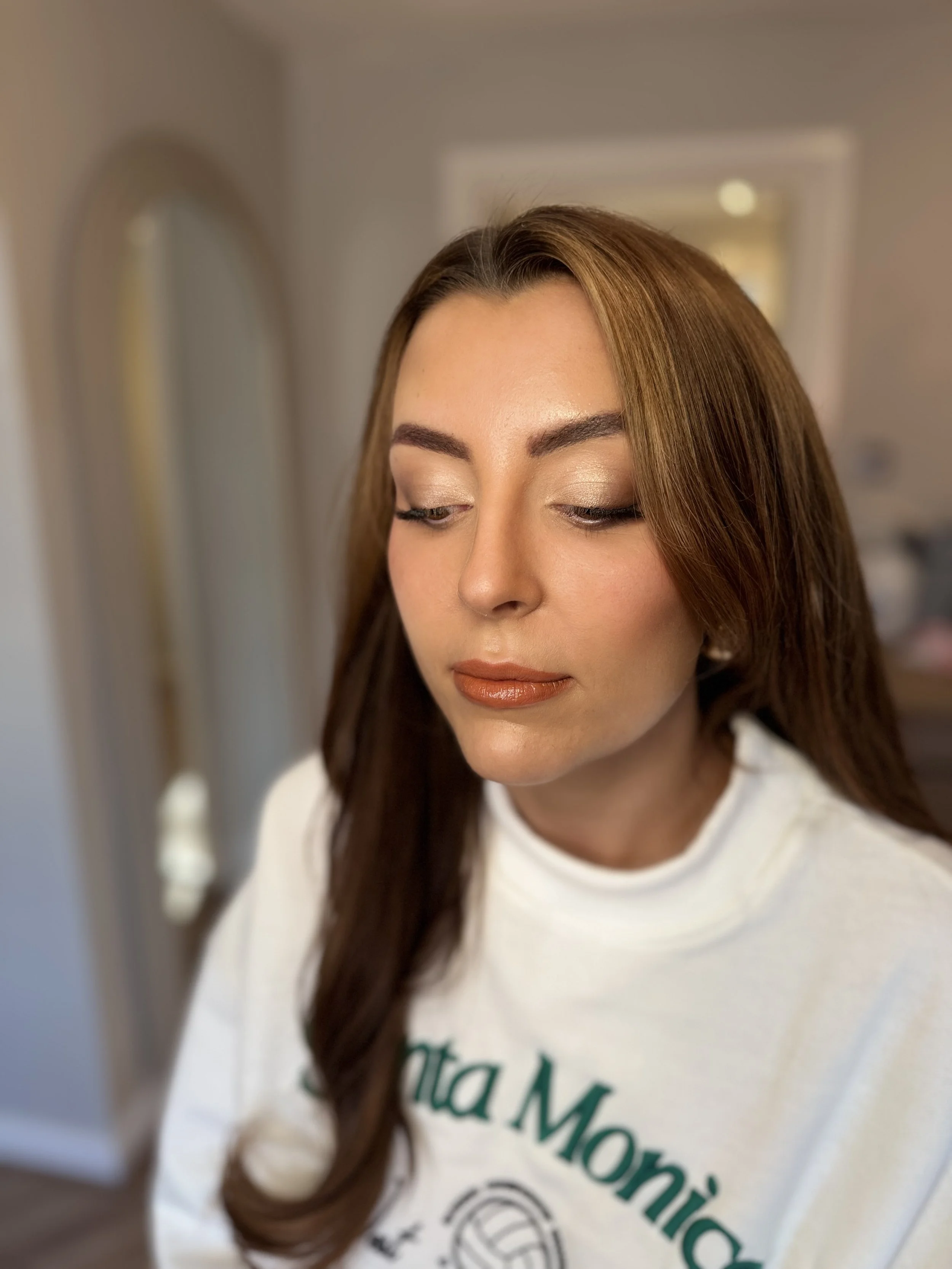 Close-up of a woman with brown hair, wearing makeup, and a white shirt, looking downward in a softly lit room.