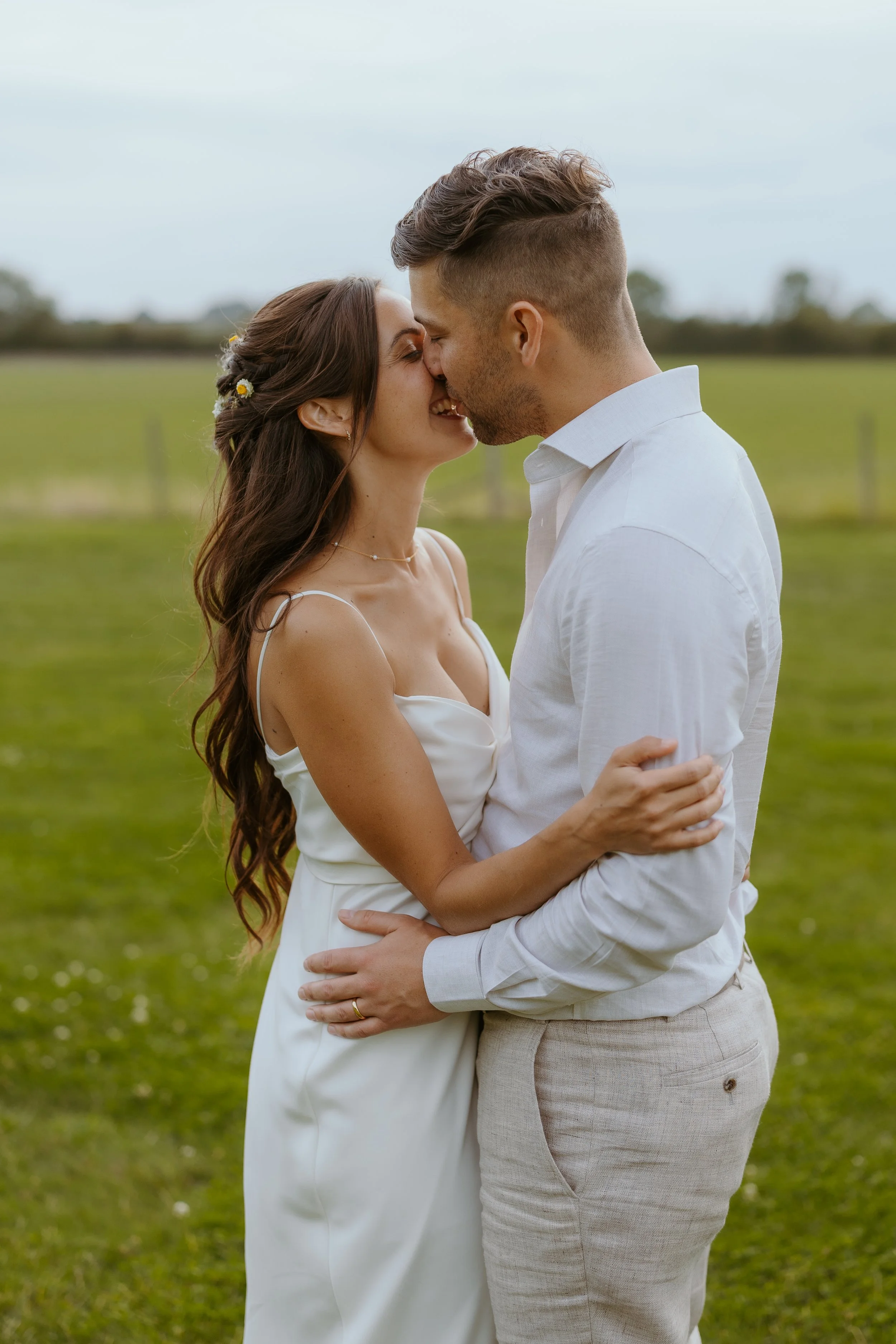 A couple dressed in white embracing outdoors on a grassy field, about to kiss, with a blurred background of trees and an overcast sky.