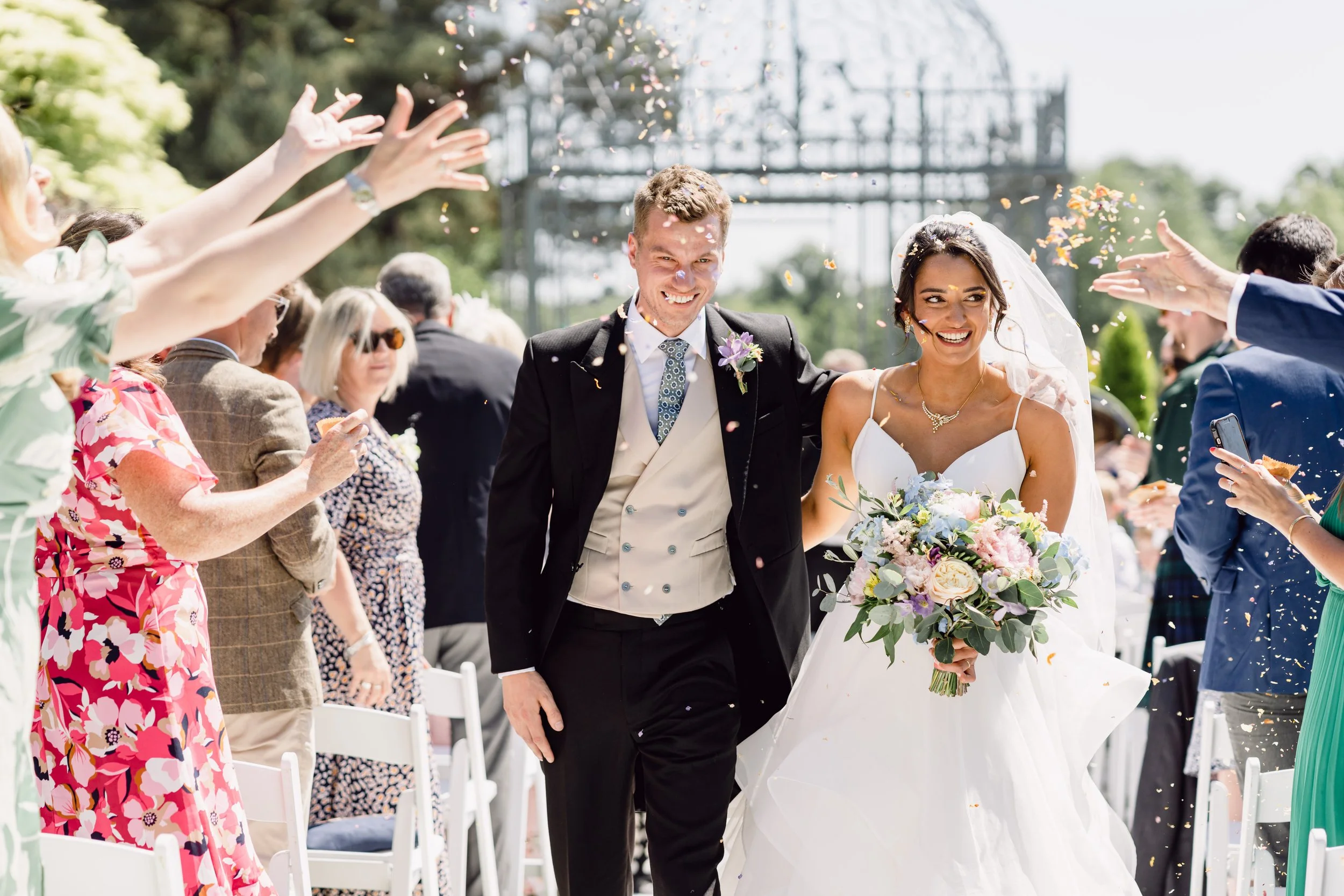 Bride and groom walking down the aisle, smiling, as guests throw confetti in an outdoor wedding ceremony.
