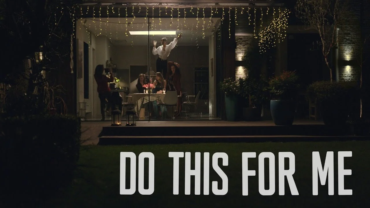 A group of women celebrating inside a house with string lights hanging outside. One woman is standing on a table dancing, while others sit or stand around the table. The scene is viewed through large glass doors, with the outdoor area lit by small ca