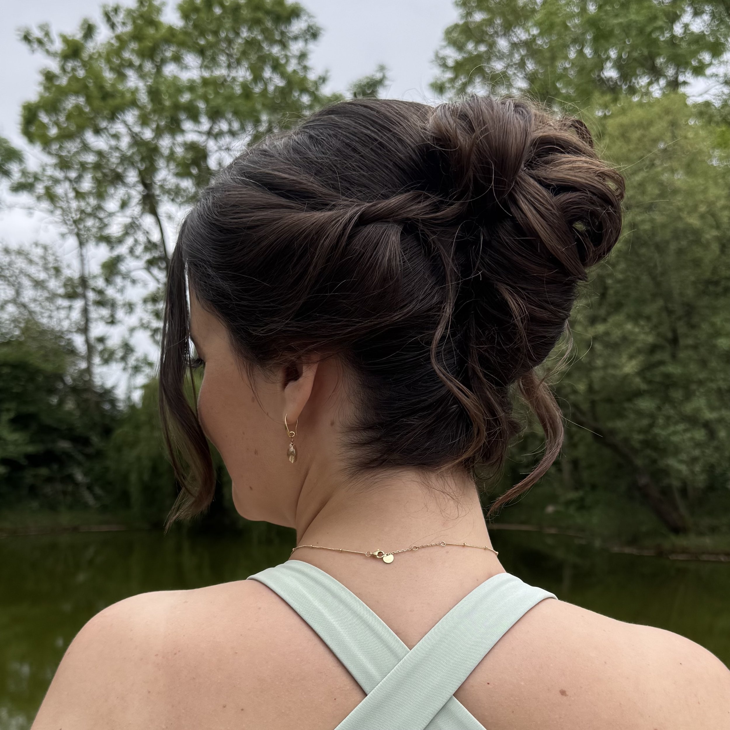 Woman with dark hair styled in an elegant updo, wearing earrings and a delicate necklace, outside near a body of water with green trees in the background.