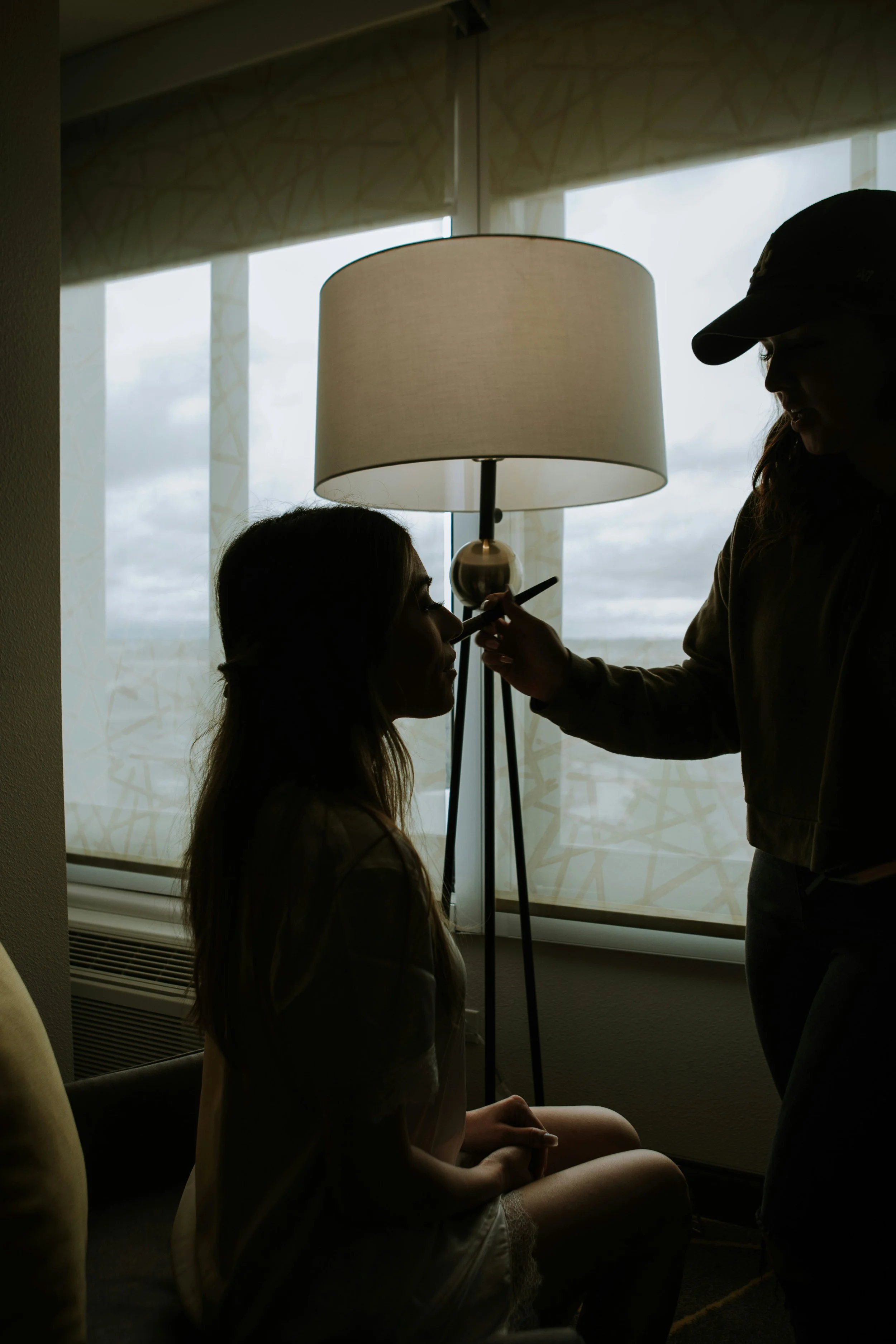 A woman sitting on a chair getting makeup applied by another woman standing beside her in front of a window with blinds.