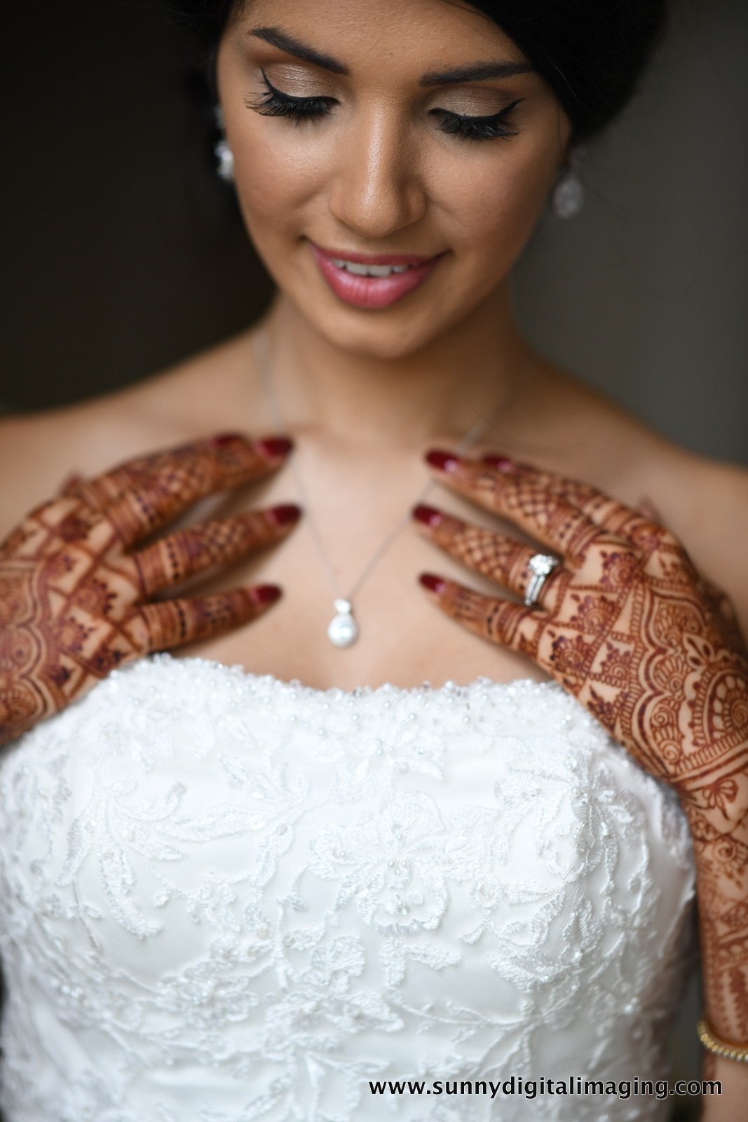 A bride smiling with henna decorated hands, wearing a white wedding dress, jewelry, and a necklace with a pearl pendant.