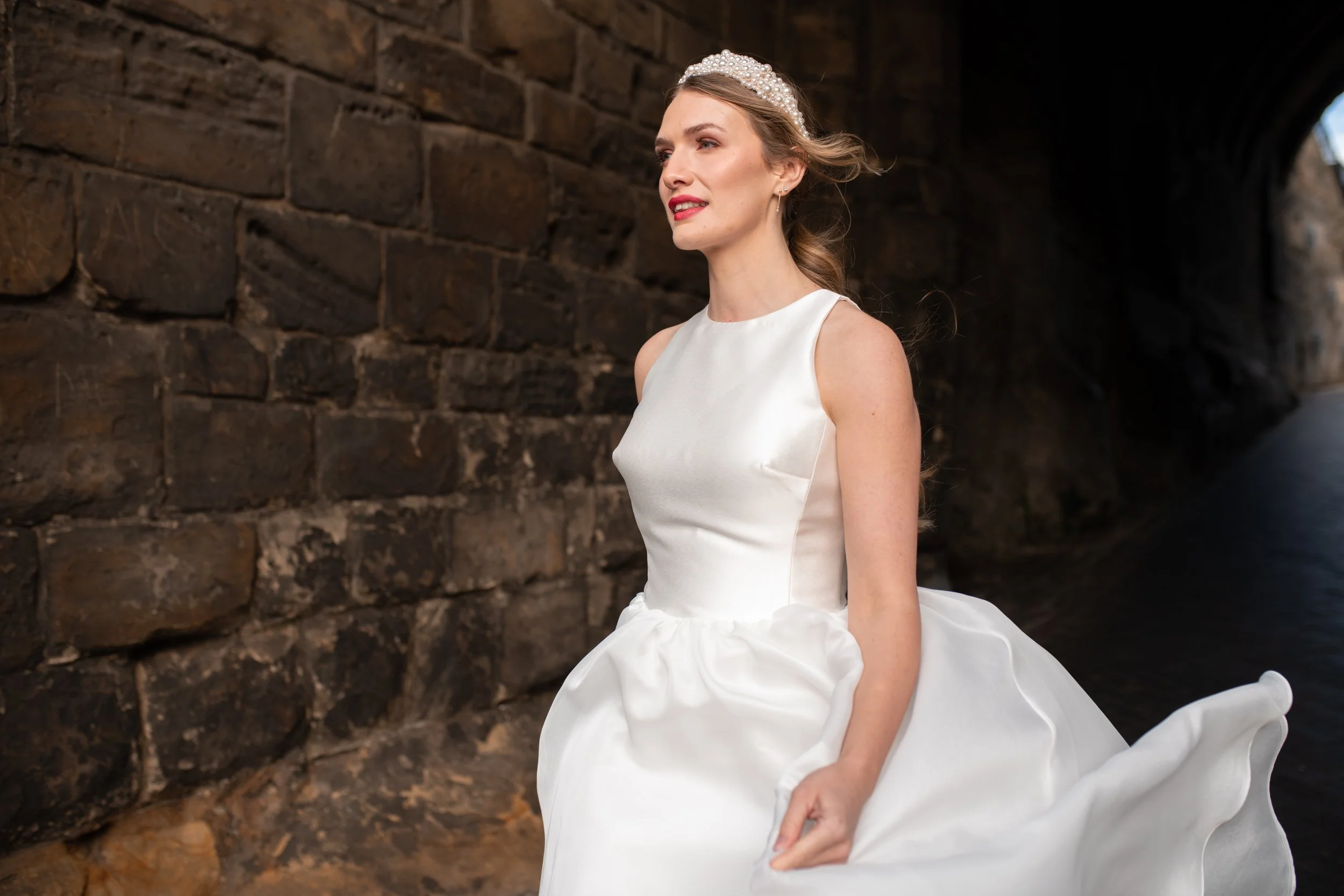 A woman in a bridal gown standing outdoors near a stone wall, with a tunnel in the background.