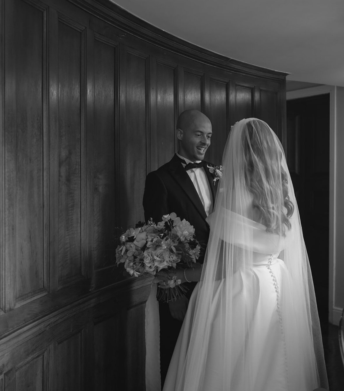 A black and white photo of a bride and groom facing each other indoors. The groom, in a tuxedo, smiling at the bride who is holding a wedding bouquet and wearing a wedding dress with a long veil.