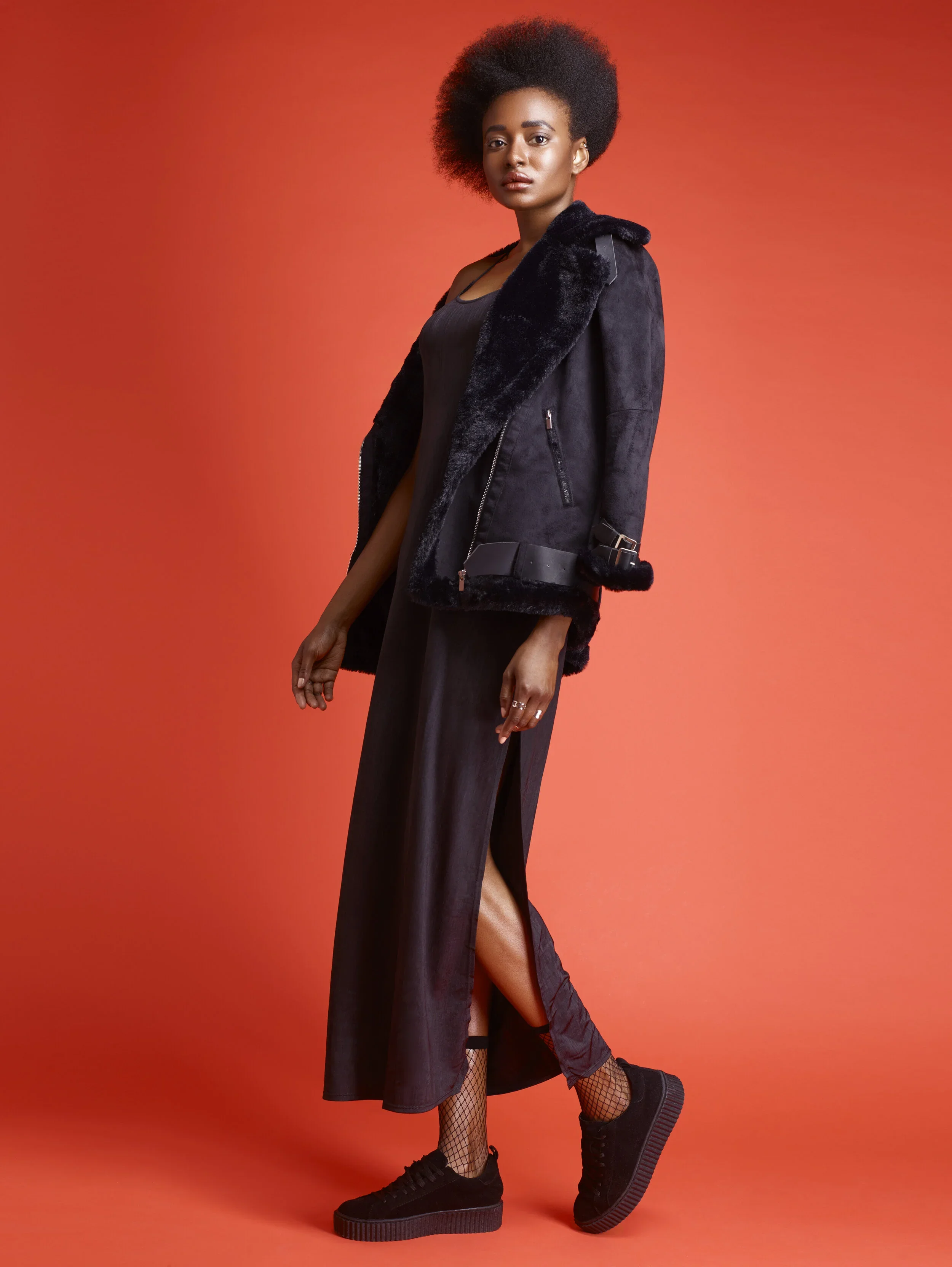A woman with an afro hairstyle wearing a black leather jacket with a shearling collar, a black dress with a side slit, fishnet socks, and black sneakers, standing against a reddish-orange background.